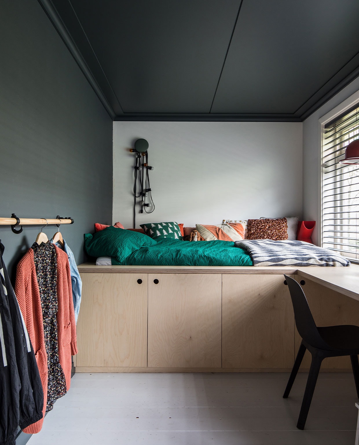 Bed, kasten en bureau zelf ontworpen en gemaakt van multiplex. Kledingrail van oude steigerdelen. Op muur en plafond Full Marble, op de achterwand wit RAL 9016 en op de vloer trappenlak van Flexa. Vlinderjaloezieën Bece raamdecoratie, Hubo. Stoel Odger Ikea. Groene knop Dots Muuto, Flinders. Lichtstreng Kwantum. Dekbed-overtrek Hema. Sprei Ikea. Kussen rood, beige, bruin-roze en pluchen beagle Ikea. Groen kussen Ferm Living. Rood kussen La Redoute. Oranje-beige kussen HKliving. Rode lamp kringloop, design hanglamp Jieldé brocante markt Lille.