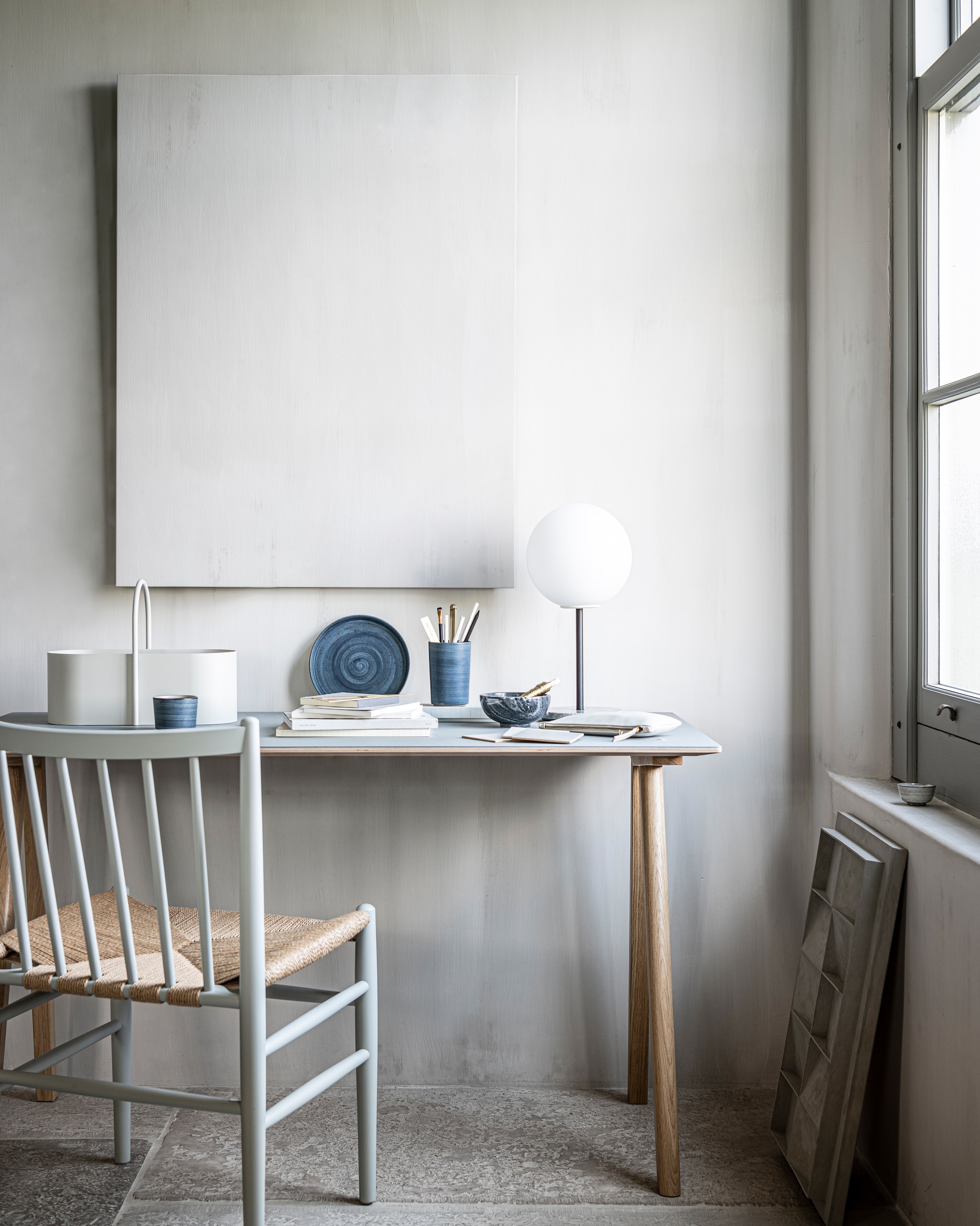 Op de wand kalkverf Old Flax van Pure &amp; Original 1 liter € 40,75 (pure-original). Bureau CPH90 van Hay 130×65 cm € 699,-, tafellamp TR Bulb van Menu met grijs marmeren plateau, zwart metalen statief en opaal glas bol € 158,99, grijze toolbox Grib van Ferm Living € 65,- (vtwonen.be). Kunstwerk N°05 in kleur Stone, 60×40 cm prijs op aanvraag (atelierledition.com). Blauw servies vanaf € 88,95, houten stoel met gevlochten zitting FDB Møbler vanaf € 649,- (letoile.store). Overig privébezit.