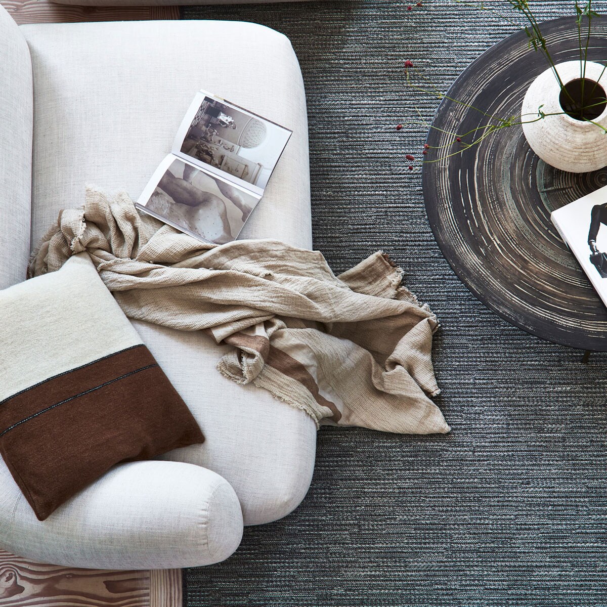 Wollen karpet Nature cord antraciet melé vanaf € 735,- M.m.v. letoile.store (bruin-wit kussen, plaid), homestede.nl (salontafel), zarahome.com (keramieken vaas op bruine salontafel, stenen vaas op bank met gevlochten zitting, houten opbergbox, slippers), homestede.nl (houten bank met gevlochten zitting), vantil.nl (voetenbank), sissy-boy.com (kom). Overig privébezit.
