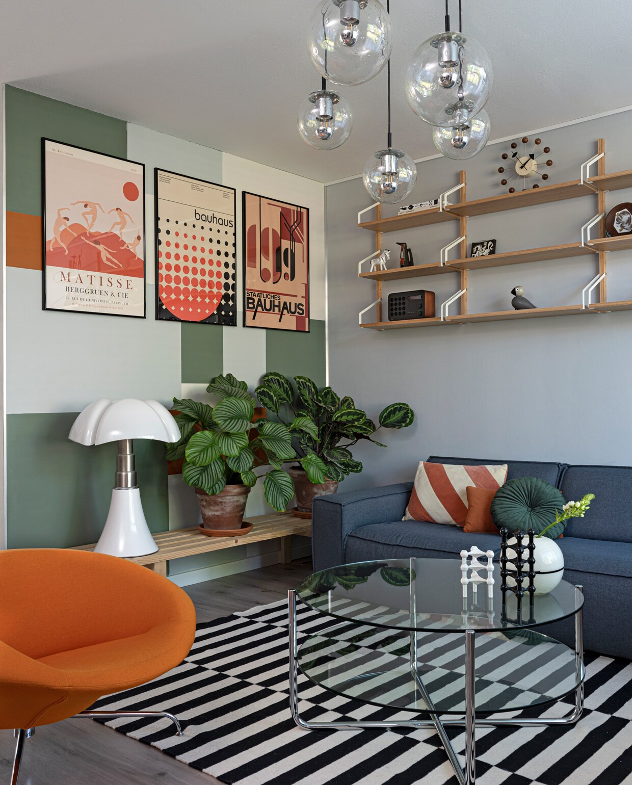 Hanglampen Raak Amsterdam via Marktplaats. Posters pstr.studio, lijsten Yourdecoration. Muren leemgroen en zinkgrijs Karwei, Indian spice Flexa. Plankjes Svalnäs Ikea. Met daarop: radio Eames, houten figuren Kay Bojesen (via Flinders), Ravelli keramiek rommelmarkt. Plantenpotten Intratuin, houten bankje zelf-gemaakt, tafellamp Pipistrello van Martinelli Luce via Marktplaats. Stoel Kirk van Artifort, glazen tafel Gispen via Marktplaats. Vloerkleed en vloer Ikea. Kandelaren Nagel via Catawiki, wit vaasje rommelmarkt. Bank Brooklyn Woon online, kussens Fonq, Loods 5 en Sissy-Boy Homeland.