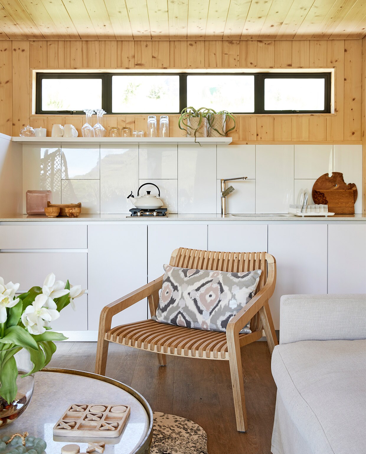Kim koos voor witte, glanzende tegels en melamine kastjes. In het blad is gerecycled glas verwerkt. Kraan Hans Grohe. Keukenblad ProQuartz. Houten fauteuil Radius Minima. Koeienhuid African Leather and Skins. Salontafel Coricraft. Overig onbekend.