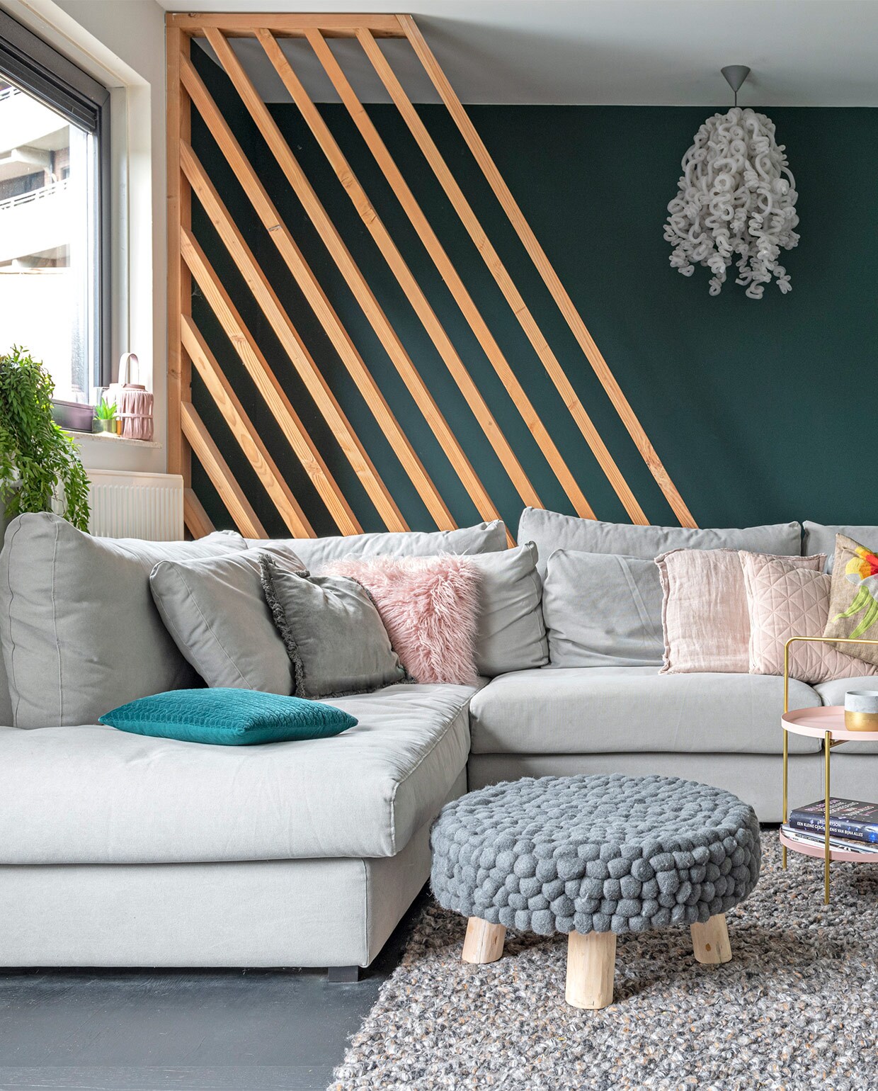 Lattenframe van douglas-hout, gemaakt door bouwbedrijf Klomp. Kleur groen op de wand Quiet Clearing van Histor. Eikenhouten vloer in blokpatroon zwart afgewerkt, vloerkleed, bank LS Napels, kussens, plaid, bijzettafel met draagbeugel, poef Loods 5. Hanglamp, merk onbekend. Bijzettafel met zwart frame Karwei. Kussen met bloem Malagoon. Groen-witte vaas vintage.