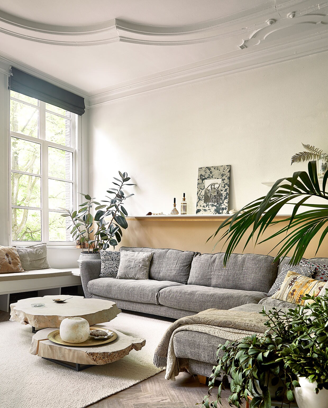 Raambekleding en vloer zaten al in het huis. Bank in vensterbank op maat gemaakt. Loungebank Seats. Kussens, plantenmand, tafels, plaid, kaarsenhouders/vazen, marmeren schaaltje en dienblad Zenza. Wollen vloerkleed Helsinki van Vloerkleden-winkel. Muurverf zelf gemengd. Bijzettafels Heerenhuis Manufactuur, fossiel van schelp, koperen schaaltje MaisonNL. Portret door dochter Naomi gezeefdrukt en geborduurd.