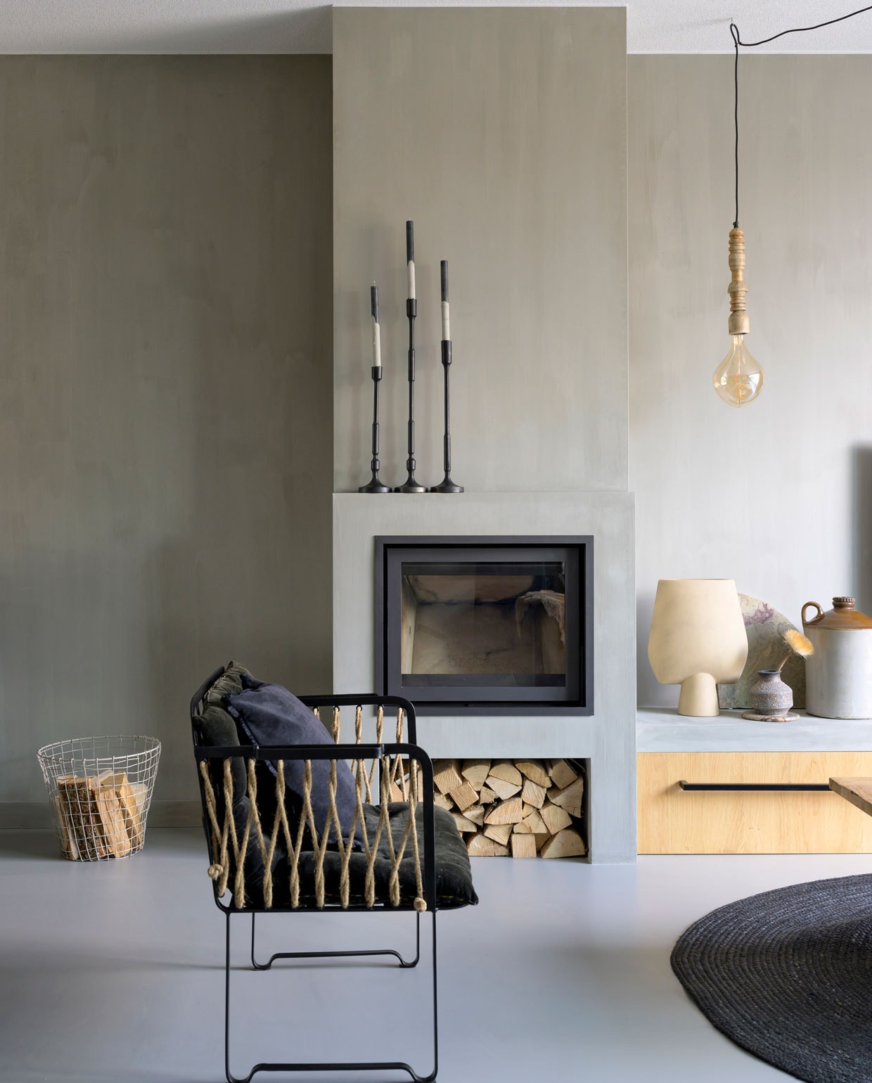 Grijze woonkamer met open haard | Houtkachel Tibas. Wandbank met lade maatwerk. Draadmand Mica Decorations. Kandelaars, hanglamp Casa Vivante. Peertje Hornbach. Vazen Casa Gitane. Grote kruik en fauteuil Madam Stoltz. Vloerkleed Karwei. Muurverf CC020 Zinco van Carte Colori.