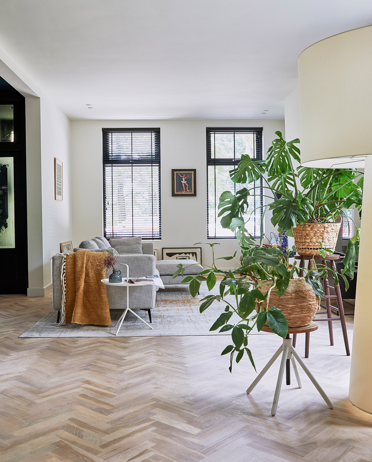 Bank Zitmaxx Wonen, vloerkleed Louis de Poortere, bijzettafel Hay, plaid Kwantum. Lage kruk Marktplaats, hoge kruk uit de inboedel van het huis. Rieten plantenmanden Xenos, schilderij op de grond Wally Elenbaas, schilderij van ballerina Herman Bieling, witte staande lamp Big Shadow van Cappellini.