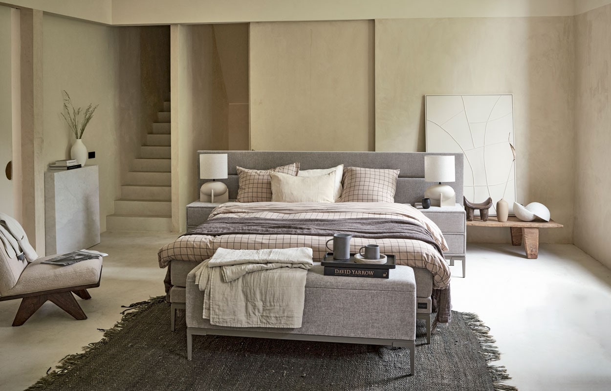 Boxspring Lifestyle by vtwonen Cinnamon Large vanaf € 1750,- (140×200 cm), nachtkastje Cardemom met twee lades, 45x45x62 cm (lxbxh) vanaf € 199,-, voetenbank Clove 140x33x57 cm (lxbxh) vanaf € 339,-, verkrijgbaar met verschillende stoffen en kleuren. Hier uitgevoerd met stof Mirage Taupe, metalen frame in kleur Champagne en poot Track. M.m.v. v-i-v.nl (schilderij), allestedlund.com (fauteuil en schemerlampen), household­hardware.nl (witte vaas), nordal.eu (linnen plaid grijs, kan en beker, marmeren schaal, linnen kussen), hm.com (linnen plaid zand op bankje), anoukb.com (vloerkleed, houten tafel, dienblad, bolvormige lamp), vtwonen.nl (vaas zandkleur, beddengoed Grid kleur zand uit de vtwonen collectie).