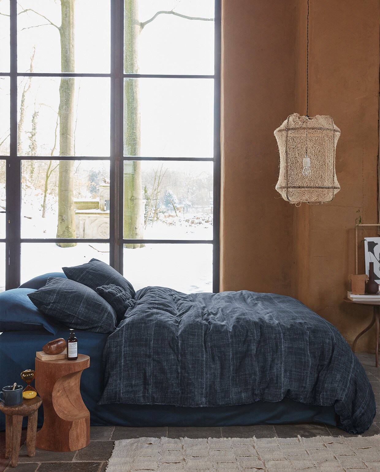 Kussen Tufted van katoen canvas, donkerblauw, 30×50 cm € 29,95. Dekbedovertrek Vintage Indigo van katoen, donkerblauw, 140×200/220 cm € 99,95, 200×200/220 cm € 189,95, 240×200/220 cm € 199,95.M.m.v. polspotten.nl (houten bijzettafel, donkerbruine vaas), studio-yen.com (vloerkleed, oude houten bijzettafel, houten steenvorm, keramieken pot), kasstoor.nl (ronde tafel driepoot), letoile.store (mok, lepel, zandloper, hanglamp, poster in lijst).Overig privébezit.