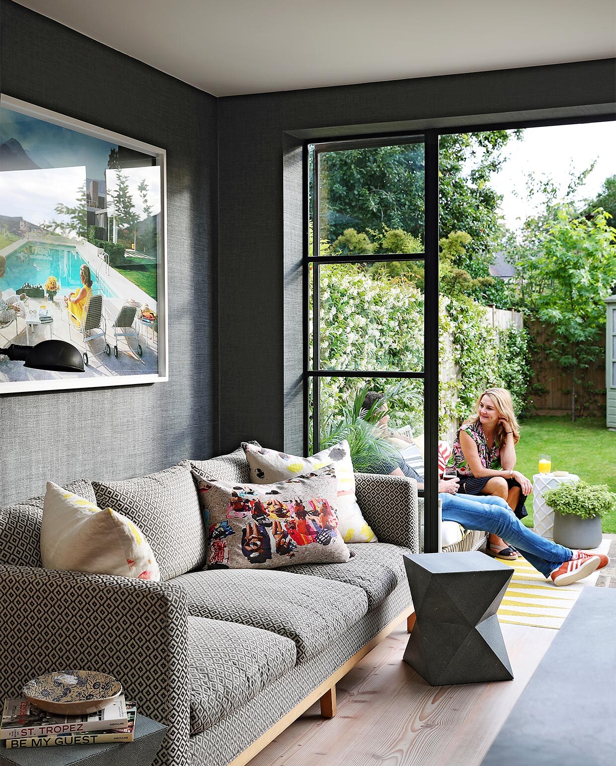 Deuren met zwart stalen kozijnen geven toegang tot de tuin. Behang Textures Végétales van Élitis in houtskooltint. Fotokunst Poolside Gossip van Slim Aaron. Bank en bijzettafels Cécile &amp; Boyd. Vierkante kussens bekleed met stof Arty van Pierre Frey, rechthoekig kussen Alexandre M-S. Overig onbekend.