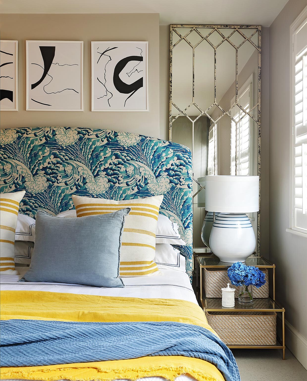 Muurverf Cornforth White van Farrow &amp; Ball. Het hoofdbord is bekleed met stof The Wave in de kleur mineral van Linwood Fabric. Beddengoed Granny Goose. Kussens met gele streep van stof Marina in kleur pastis van Lelièvre. Vaalblauwe linnen kussens lokaal gekocht. Gele en blauwe plaids H&amp;M Home. Spiegel Block &amp; Chisel. Tafellamp Gioalo van OKA. Bankje bekleed met stof van Mavromac. Kunst in lijsten gekocht via Etsy.