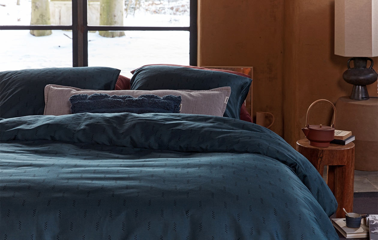 Kussen Tufted van katoen canvas, donkerblauw, 30×50 cm € 29,95. Dekbedovertrek Crops van katoensatijn, donkerblauw, 140×200/220 cm € 79,95, 200×200/220 cm € 149,95, 240×200/220 cm € 159,95 (ook in donkerrood).M.m.v. polspotten.nl (houten kruk), letoile.store (theepot, mok, lepel), loods5.nl/design (papier-maché bijzettafel in aardetint, tafellamp). Overig privébezit.
