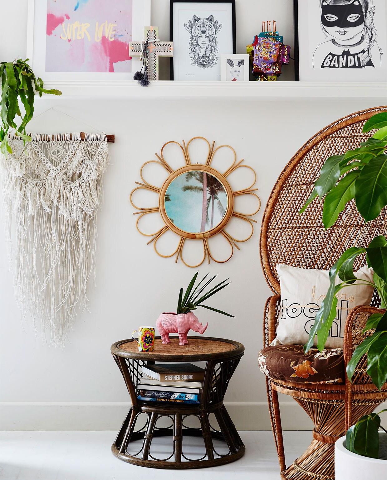 Macramé wandhanger Family Threds. Rotan spiegel @downtothewoods. Bijzettafel en stoel gevonden op straat, wit kussen Kawaiian Lion. Roze neushoornplantenpot Rice dk. Op plank: keramieken kruis met hanger Ahoy Trader, print Bandit Girl Mini &amp; Maximus. Plant in pot Olive Green Plant &amp; Prop. Overig onbekend.