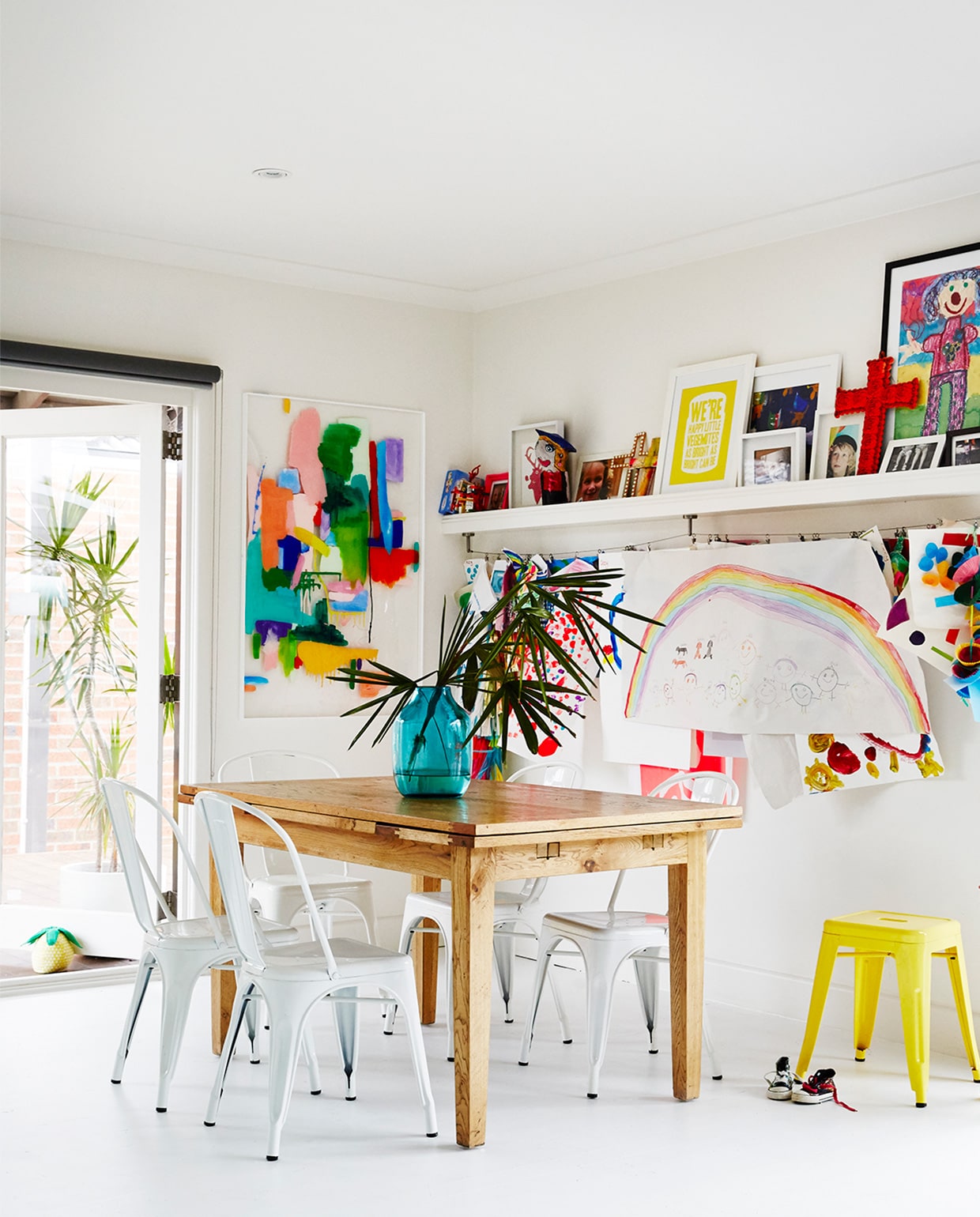 Kleurrijk schilderij op plexiglas van Kristen Daniels. Uitschuifbare eettafel vintage. Witte, metalen terrasstoelen en gele kruk Tolix. Op de wandplank en aan het koord eronder worden de tekeningen van de kinderen uitgestald.