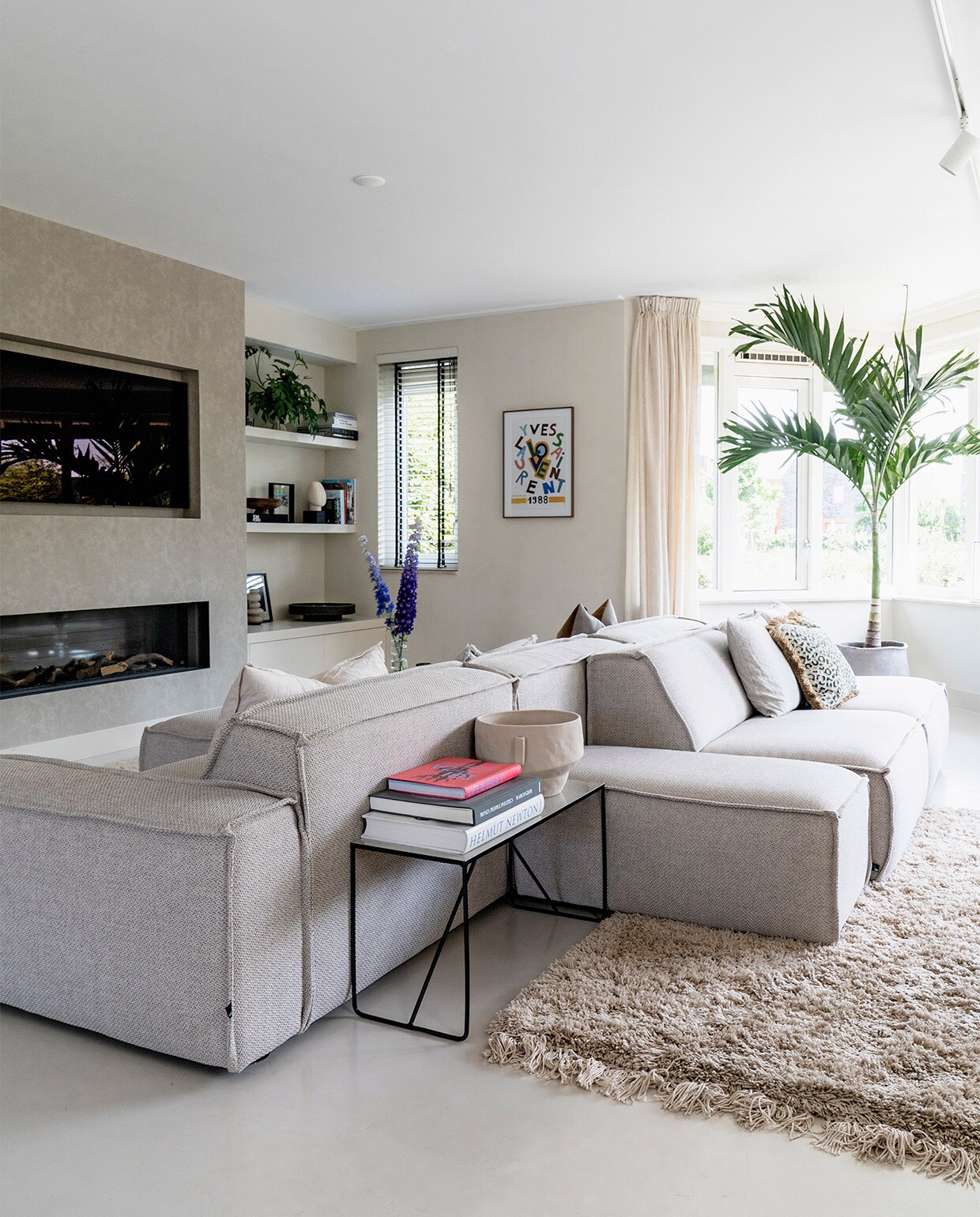 Vloer Tav Microcement. Modulaire bank Edge van Fest Amsterdam. Bijzettafel Broste Copenhagen. Vaas Zara Home. Televisie LG. Haard Dru. Poster aan de wand vintage. Kussens onder andere Nox en Loft. Plant Veitchia merrillii. Vloerkleed Westwing.