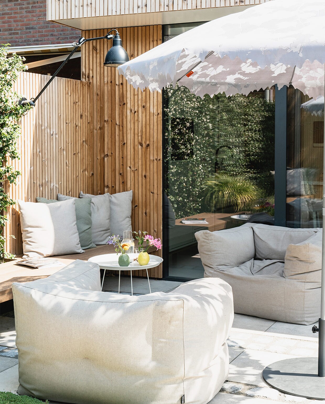 Latten rondom het huis thermo pine design rabat. Tuinstoelen Bryck. Op maat gemaakte kussens Sunbrella. Bijzettafel Fatboy. Parasol Droog. Hanglamp DCW Editions. Gele en groene vaas bloemenwinkel Jasmijn.