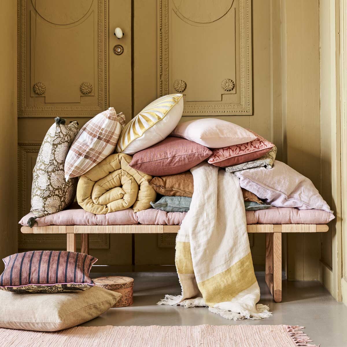 Bankje BM0488 van Børge Mogensen &amp; Carl Hansen, 138x46x34,5 cm (bxdxh) € 1440,-, geruit kussen € 49,90, plaid met streep van Libeco,110×180 cm € 122,-, donkerroze kussen, 40×80 cm € 79,90, klein roze kussen, 30×40 cm € 44,90, (letoile.store). Roze vloerkleed Chindi Mat Rose, 60×90 cm € 20,-, kussen linksonder op de grond, 50×50 cm € 45,-, kussen met print links Shimla Olive, 50×50 cm € 47,-, geel matras Mirra Lemon Curd, 55×150 cm € 55,-, roze matras Mirra Rose, 55×150 cm € 55,-, rond kussen met geel borduurwerk € 95,-, groen kussen, 50×50 cm € 47,-, oker kussen, 50×50 cm € 47,-, lila kussen, 50×50 cm € 47,-, groen tafelkleed met print, 150×250 cm € 100,-, roze kussen met borduurwerk bloemen, 30×40 cm € 53,75, alles van Bungalow (scandicliving.nl). Kussen met strepen linksonder Doris van HKliving, 30×40 cm € 57,95 (hkliving.nl). Doosje € 3,- (jolandelijkdewebshop.nl). Kleur op de wand vergelijkbaar met Gervase Yellow No.72 van Farrow &amp; Ball