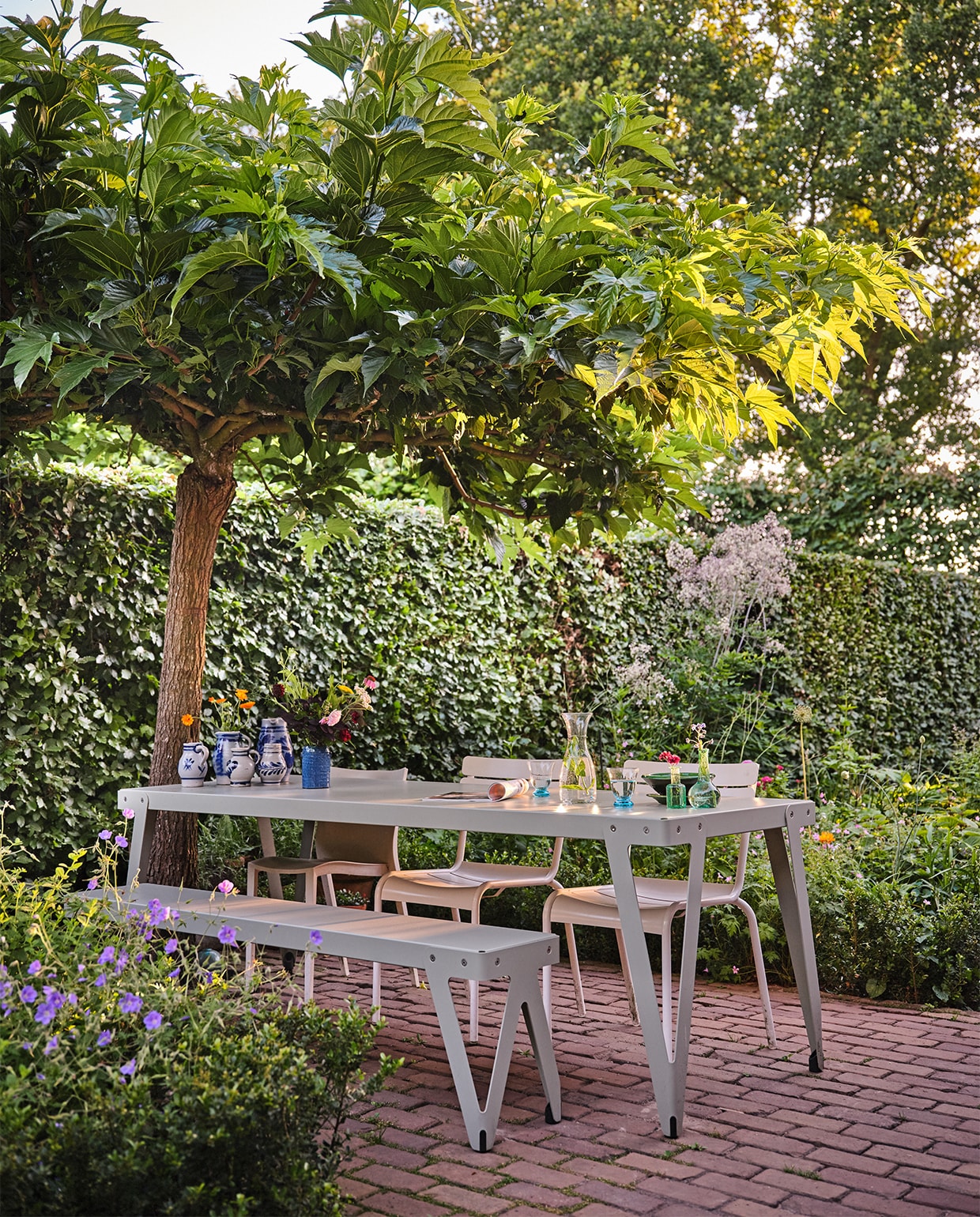 De tafel in de zijtuin. Blauwe geranium op voorgrond: Johnson’s Blue. In het vak achter is nog een wolk van de lila/roze ruit (thalictrum) zichtbaar. Tafel en bank Functionals, twee stoelen Fermob. Op de tafel: Keulse potten Marktplaats, diverse vaasjes De Weldaad, blauw vaasje Søstrene Grene. Glaswerk meegenomen uit Spanje.