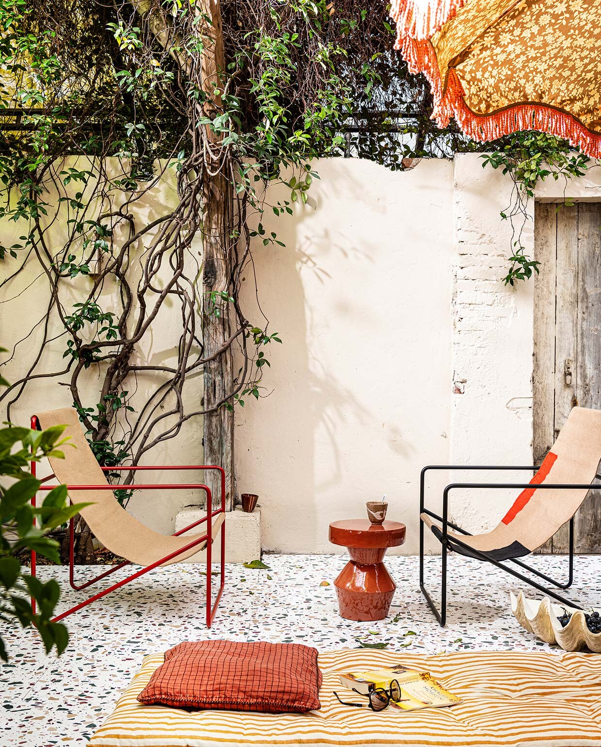 Terrazzo vloer van de buitenplaats styling Capri, 60×60 cm (lxb), voor binnen en buiten, mits onder afdak (tegelbv.com). Stoelen Desert van Ferm Living (vtwonen.nl). Rode bijzettafel (urbannatureculture.com). Gestreept dekmatras 180×70 cm (lxb) en kussenhoes allebei Madam Stoltz (deens.nl). Polystone schelp (nordal.eu). Parasol van HKliving (vtwonen.nl). Kopjes, set van 4 stuks (hkliving.nl).