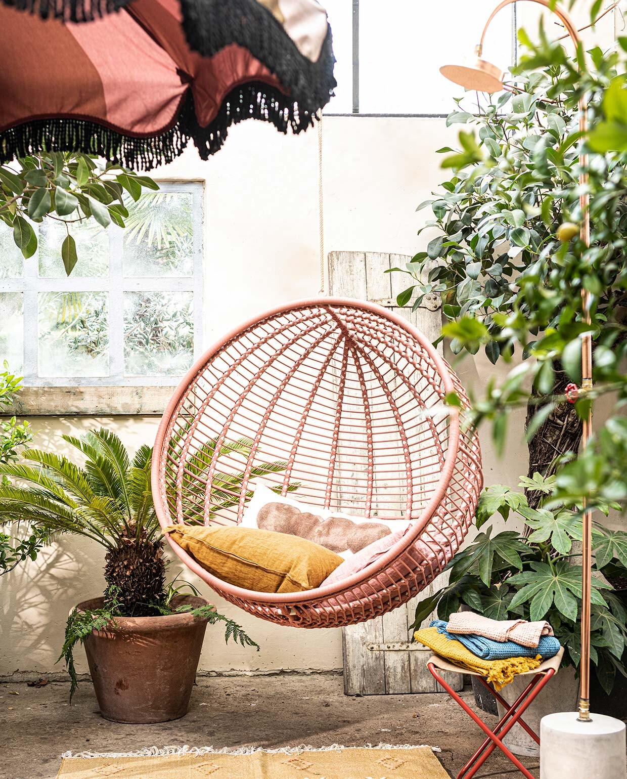 Stalen hangstoel voor buiten in zacht terra, 107×73 cm (Øxd)(kavehome.nl). Krukje Desert van Ferm Living (vtwonen.nl). Koperen buitendouche van Seletti (vtwonen.nl). Parasol van HKliving (vtwonen.nl). Langwerpig gestipt kussen (urbannatureculture.com ). Linnen kussens, wafeldoeken (bungalow.dk). Vloerkleed van Madam Stoltz (deens.nl). Plant en pot privébezit.