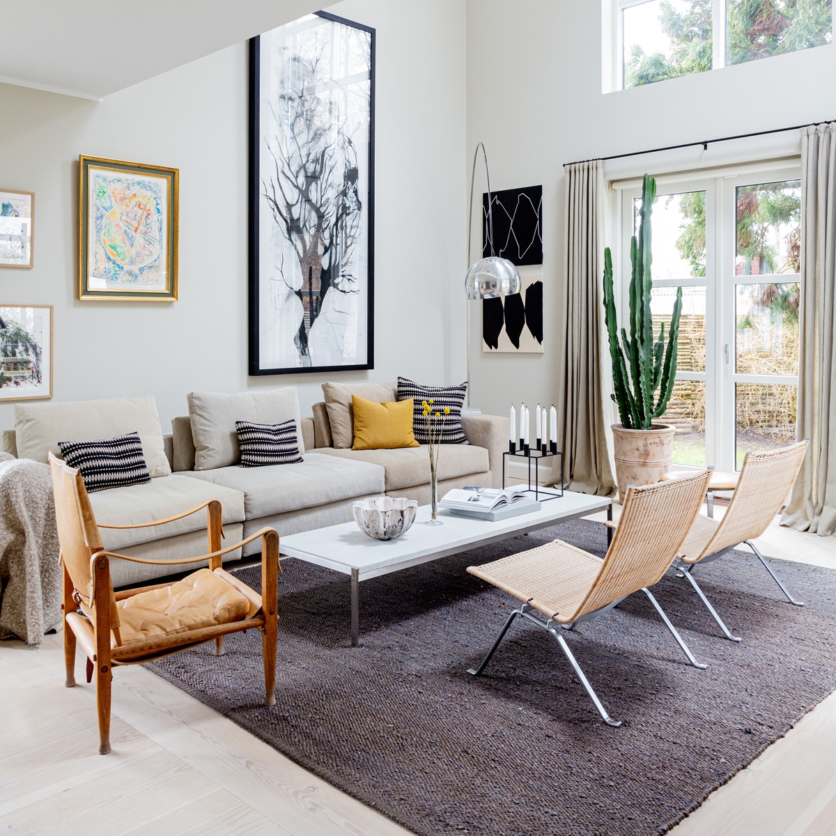 Muurverf Shaded White vanFarrow &amp; Ball. Bank Eilersen. KussensTapet-Cafe. Koffietafel PK62 en rotanstoelen PK22 van Poul Kjærholm voorFritz Hansen. Leren stoel Kaare Klintvoor Carl Hansen &amp; Son. VloerlampArco van Flos. Schaal gemaakt dooreen vriendin. KaarsenstandaardMogens Lassen. Schilderijen van linksnaar rechts Carsten Dahl (twee maal),Eigill Jacobsen, Cathrine RabenDavidsen. Grafische kunstwerkenUlla Pedersen. Vloerkleed Tine K Home.