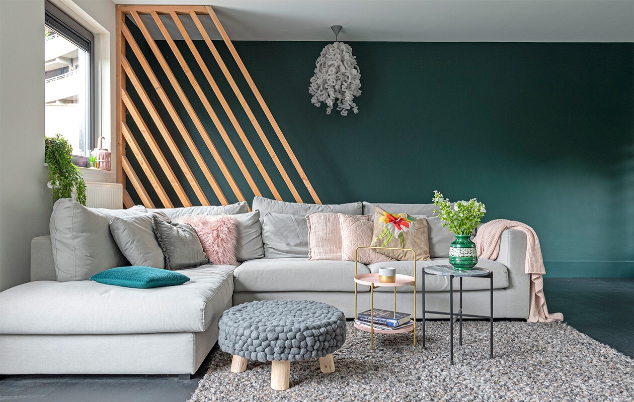 Lattenframe van douglas-hout, gemaakt door bouwbedrijf Klomp. Kleur groen op de wand Quiet Clearing van Histor. Eikenhouten vloer in blokpatroon zwart afgewerkt, vloerkleed, bank LS Napels, kussens, plaid, bijzettafel met draagbeugel, poef Loods 5. Hanglamp, merk onbekend. Bijzettafel met zwart frame Karwei. Kussen met bloem Malagoon. Groen-witte vaas vintage.