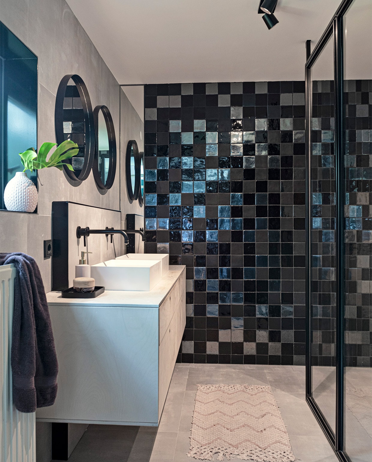 Grijze tegels op vloer en wand Spotlight van Villeroy &amp; Boch. Tegels achterwand Malaga in zwart-mat, zwart-glans en metallic Cottoceramix. Douchewand op maat gemaakt. Plafondspot Epic van Maretti. Composiet wasbak Dogma van Bliss Elements. Wandkranen uit de T-mix serie van Bongio. Wastafel van berken-multiplex door Leurs Interieurbouw. Spiegels Kwantum. Tray Zara Home. Badmat, handdoek, vaas en zeeppompje herkomst onbekend