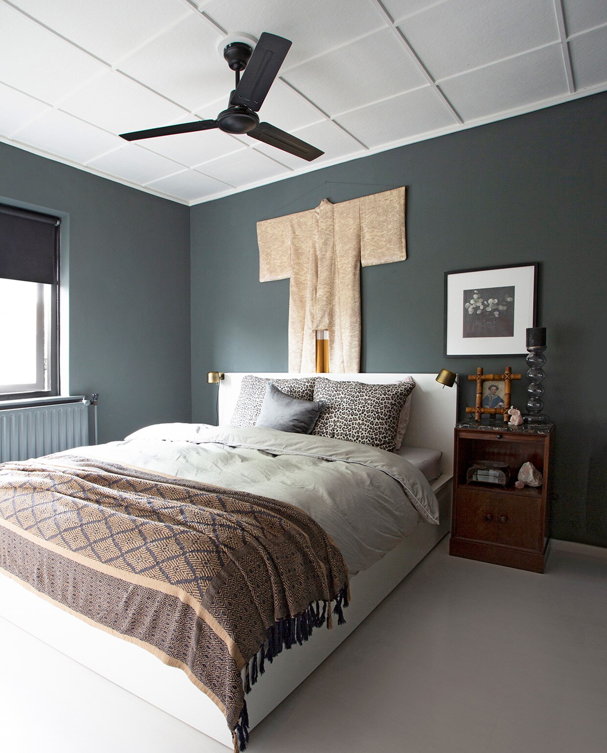 Muurverf gobi-grijs Perfection via Praxis. Bed Ikea. Vloer laminaat bouwmarkt. Ventilator Amazon. Lakens H&amp;M Home en Essenza Home. Leopard kussen Essenza Home. Kimono van de Noordermarkt in Amsterdam opgehangen aan bezemsteel. Gordijnen, nachtlamp, fotolijst, glazen kandelaar Ikea. Bamboe fotolijst antiekmarkt. Nachtkastje kringloopwinkel.