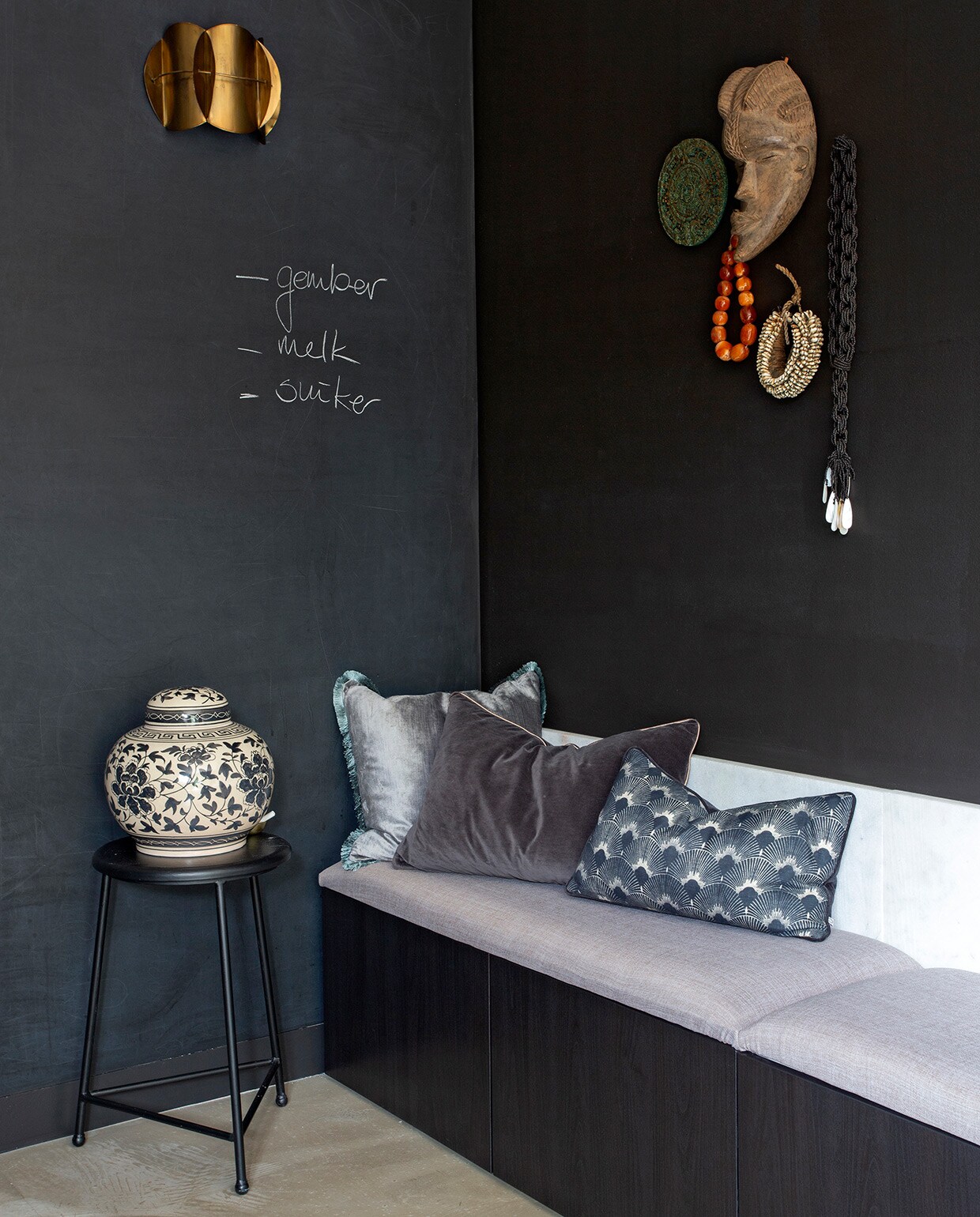Muurverf Raaf Histor. Bank van Bestå element Ikea. Zitkussens zelfgemaakt. Kussen blauwgrijs en antraciet H&amp;M Home. Kussen met waaier House Doctor. Vaas met deksel Ibiza Outdoor. Wandlamp HomeStock. Zwarte kruk, decoratiebord, masker, schelpenketting, zwart-witte kralenketting kringloopwinkel.