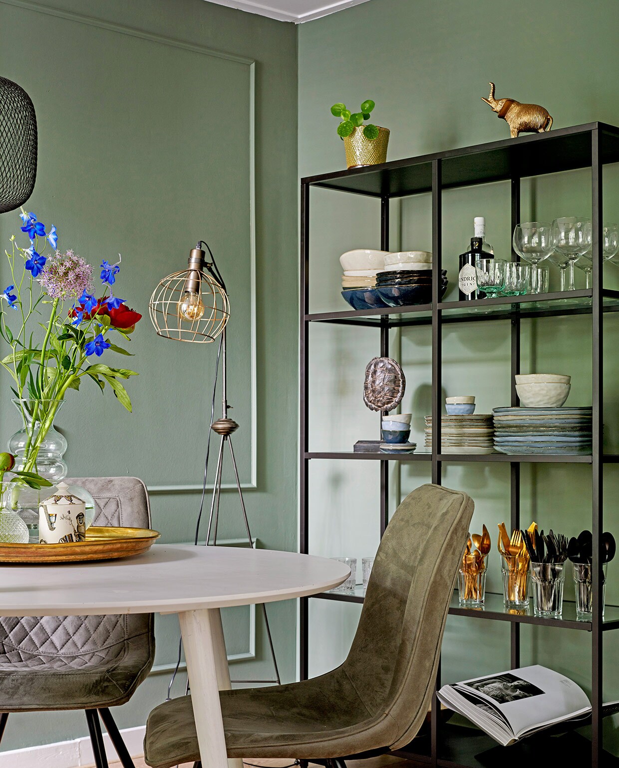 Tafel Made. Stoelen Zitmaxx. Hanglamp Ikea. Dienblad op tafel herkomst onbekend, geurkaars in potje Fornasetti, glazen vaas Living-in By Romy’s. Staande lamp Karwei. Stellingkast Ikea. Plantenpot De Bloemerije. Olifantenbeeldje en goudkleurig bestek Action. Servies Casa. Schildpaddenschild Living-in By Romy’s.