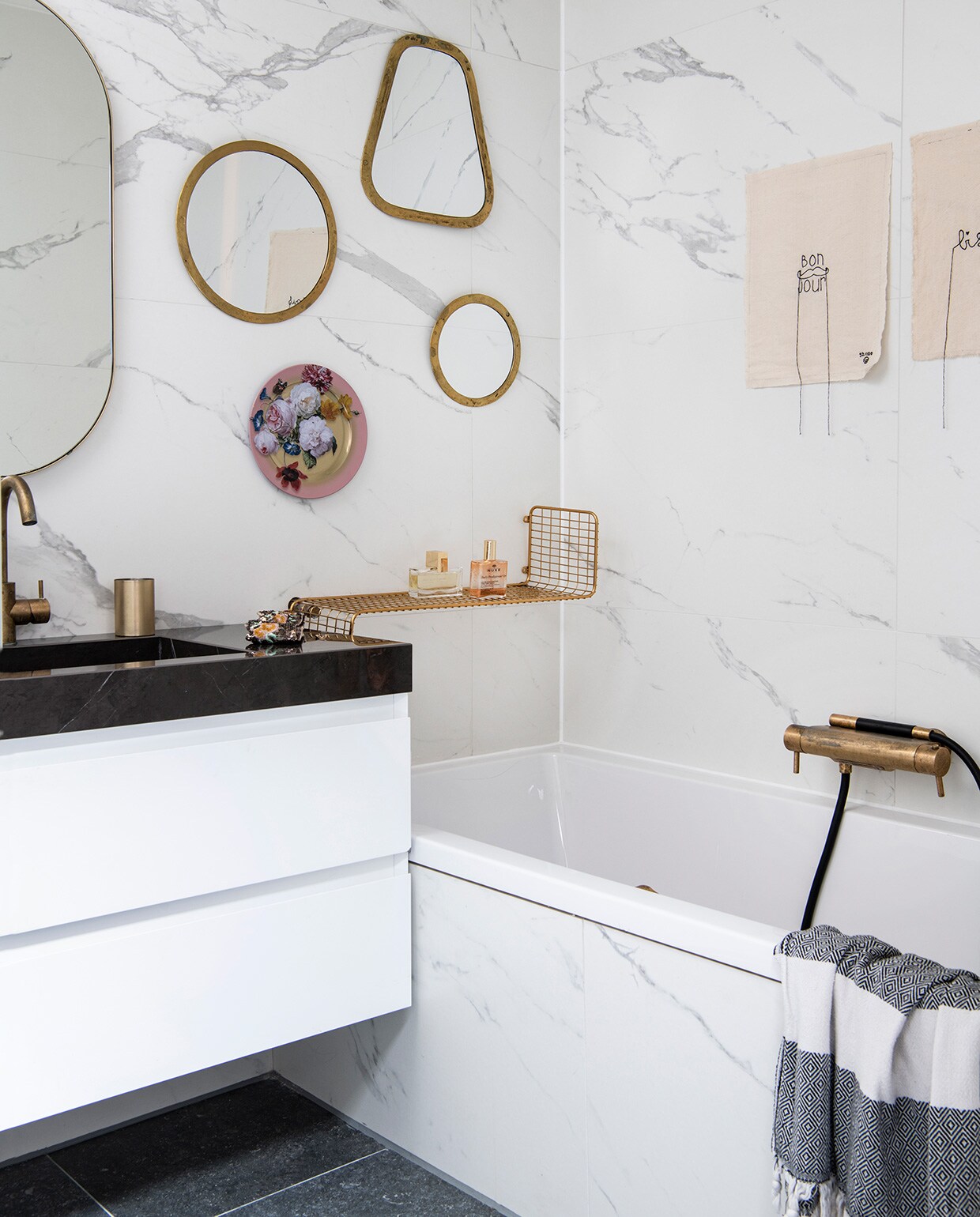 Badkamer en tegels via Ton Scholten Tegelhandel, kranen van Hotbath, messing beker uit India, grote spiegel van Made. Wandspiegels van Household Hardware, roze gebloemd bord van Loods 5, messing rek van Ikea. Hammamdoek over badrand van El Ramla Hamra. Kunstwerkjes met stikwerk boven het bad van Lemonwise.