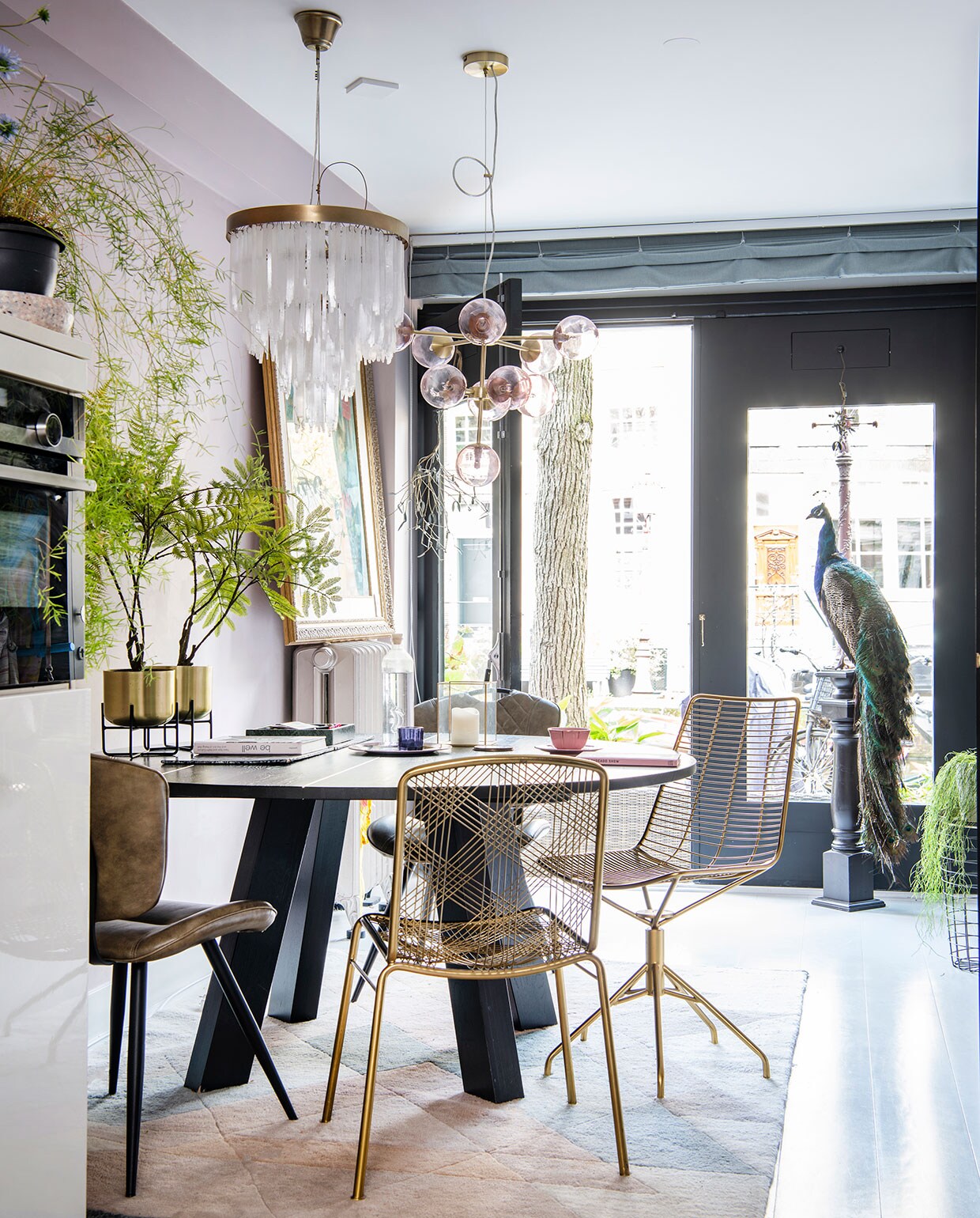 Ronde tafel van Leeelijk, leren stoelen van Wonen met Lef, de messing stoelen zijn van Zenza. Plantenhouders met messing potten Salix van Made, erin zijdeplanten van Silk-ka. Hanglamp met bollen van Made, lamp met hangende glazen pegels van Zenza. Vloerkleed van Rozenkelim, opgezette pauw van Taxidermy Amsterdam. Fles op tafel van Regalo Gallery, glas en roze kop-en-schotel van Anthropologie, blauwe waxinelichthouder van Zara Home. Het schilderij op de radiator is een souvenir uit Bali.