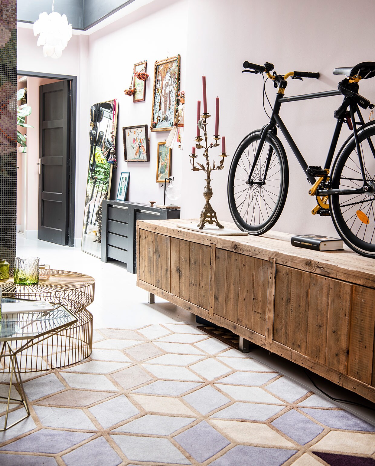 Op de wand krijtverf van Dubbelleven in de kleur Zoet. Houten dressoir van Leeelijk, fiets van Made, net als de spiegel links. Vijfarmige (antieke) kandelaar uit Puglia. Witte hanglamp van Zenza. Geborduurd schilderij van de Noordermarkt, andere schilderijen privébezit, prenten en handgemaakte kettingen uit India, antieke Chinese pop uit Parijs. Kleed van Rozenkelim. Tafel met glazen blad van L’Etoile, salontafel van draadstaal van Zara Home.