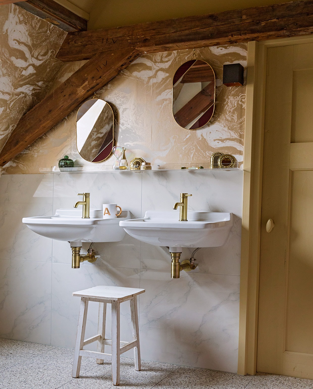 Terrazzovloertegels, keramische wandtegels met marmerprint, kraan van geborsteld koper en witte kruk herkomst onbekend. Behang Marble Gold 188 van Ferm Living. Verfkleur houtwerk Bisquit van Painting The Past. Wastafels Villeroy &amp; Boch. Plexiglas wandplankjes Ikea. Ovalen spiegels &amp;Klevering. Mok met gouden oor cadeau gekregen.