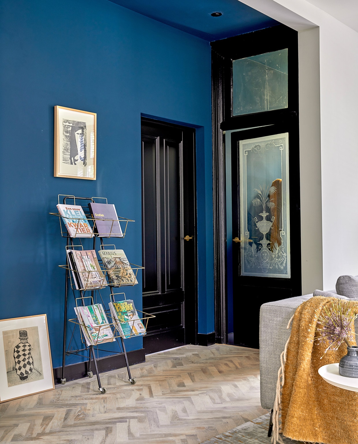 Eikenhouten visgraatvloer Balkan Wood, muurverf Haags Blauw van Farrow &amp; Ball, tijdschriftenrek kringloopwinkel, schilderij aan de muur Wally Elenbaas, schilderij op de grond Jaap Kraanen, plaid Kwantum. Verf deuren en plinten grafietzwart Ral 9011.