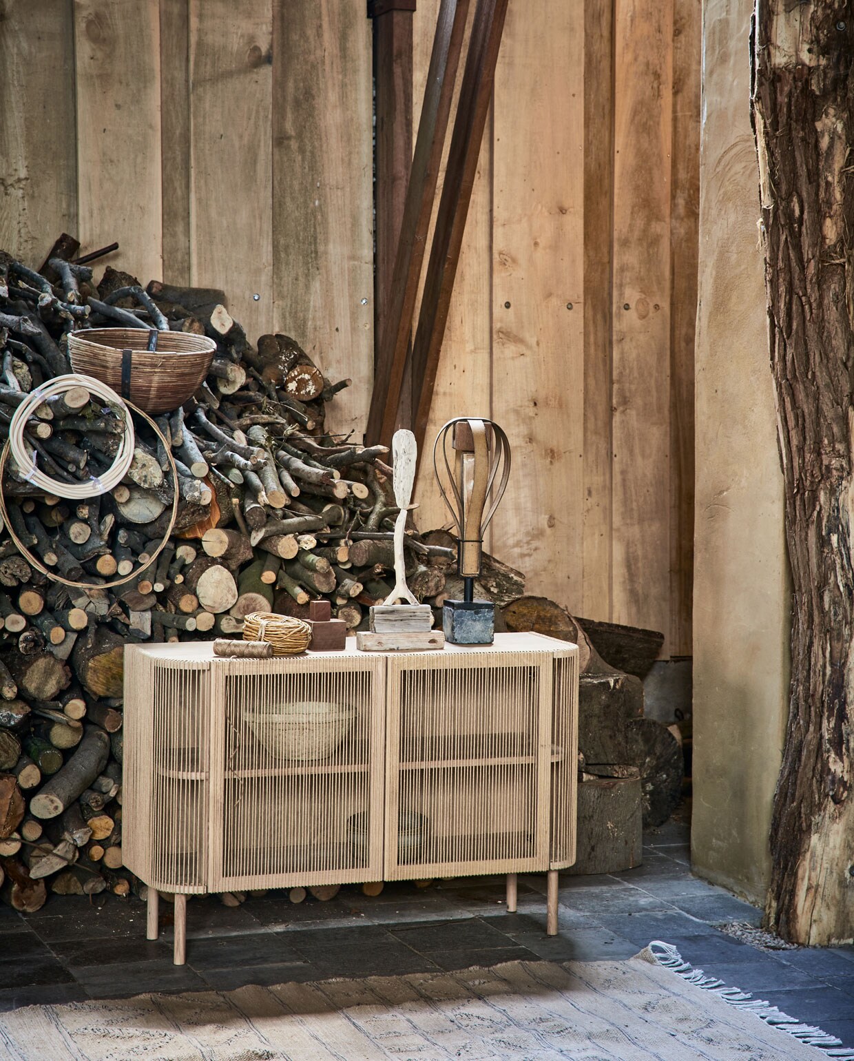 Dressoir Cord van wit geolied eiken , en touw, 120x80x40 cm (bolia.com). Kunstobjecten prijs op aanvraag (martvisser.nl). Grote rol goudkleurig koord, rotan ring prijs op aanvraag, bij elkaar gebonden spline (aangeenbrug.nl). Rotan mand, papier-maché schalen in dressoir (weldaad.com). Vloerkleed, 240×150 cm(hkliving.nl).