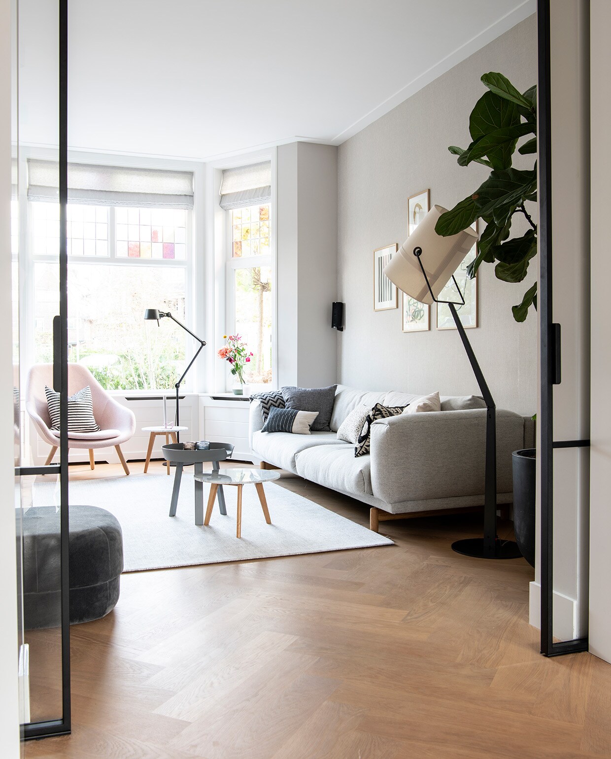 Vloer eiken. Glas in lood gemaakt in Polen. Gordijnen, wit kussen en donkergrijs kussen By Mölle. Bank en grijze bijzettafel Muuto. Plaid Elvang Denmark. Roze fauteuil Hay, gestreept kussen House Doctor. Zwarte vloerlamp Bolt van Tonone. Vloerkleed Limited Edition. Witte bijzettafel Zuiver. Poef Circus van Normann Copenhagen. Rechthoekig kussen en kussen zwart-wit patroon Mae Engelgeer. Vloerlamp witte kap Diesel. Kunst van The Poster Club in lijsten van Bolia. Zwart-wit gestreept kussen Koya Living, wit kussen met zwarte stippen Pom. Waxinelichthouders Iittala.
