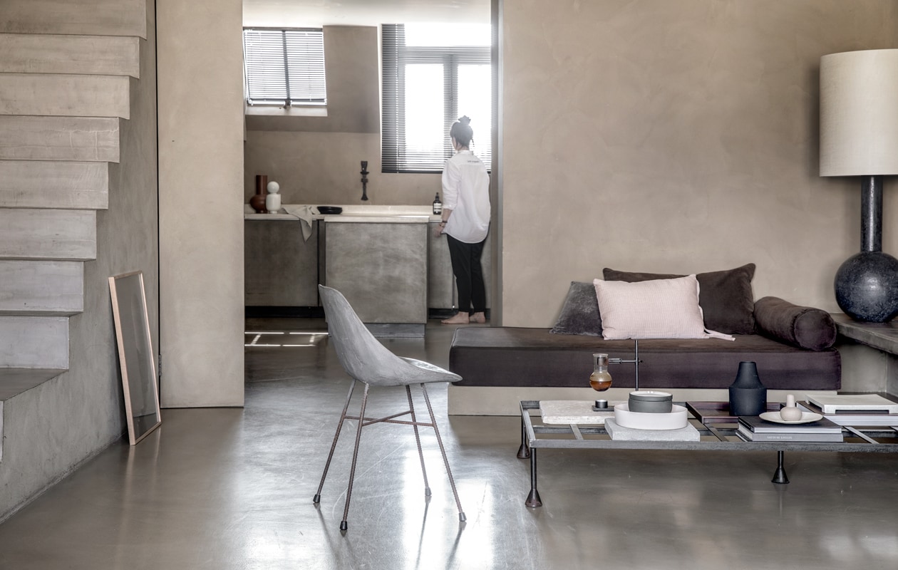 Stoel Hauteville met kuipje van beton (debetonfabriek.nl). Salontafel van gepatineerd ijzer (paullelieveld.com). Op tafel: grijze trommel van keramiek, Ivy (housedoctor.com). Zwarte vaas (loods5.nl). Klein vaasje bobbles (housedoctor.com). Sofa is eigen ontwerp; het onderstel is gemaakt van mdf, afgewerkt met beton-ciré. Erbovenop een daybedkussen – zie uitleg hiernaast. Overige kussens: grijs (loods5.nl), roze en zwart (aaimadewithlove.com). Grote lamp is vintage, gekocht bij Dolores Del Dia in Amsterdam, de kap is gemaakt door René Houben (renehouben.com). Lijst op de grond is van massief eiken 50×70 cm (papercollective.com). Op het aanrecht: vaas (loods5.nl). Fles met ronde dop Muses Talia (fermliving.com). Bord (couleurlocale.eu). Keukendoek (housedoctor.com). Zeeppompje van Skins Cosmetics (skins.nl).