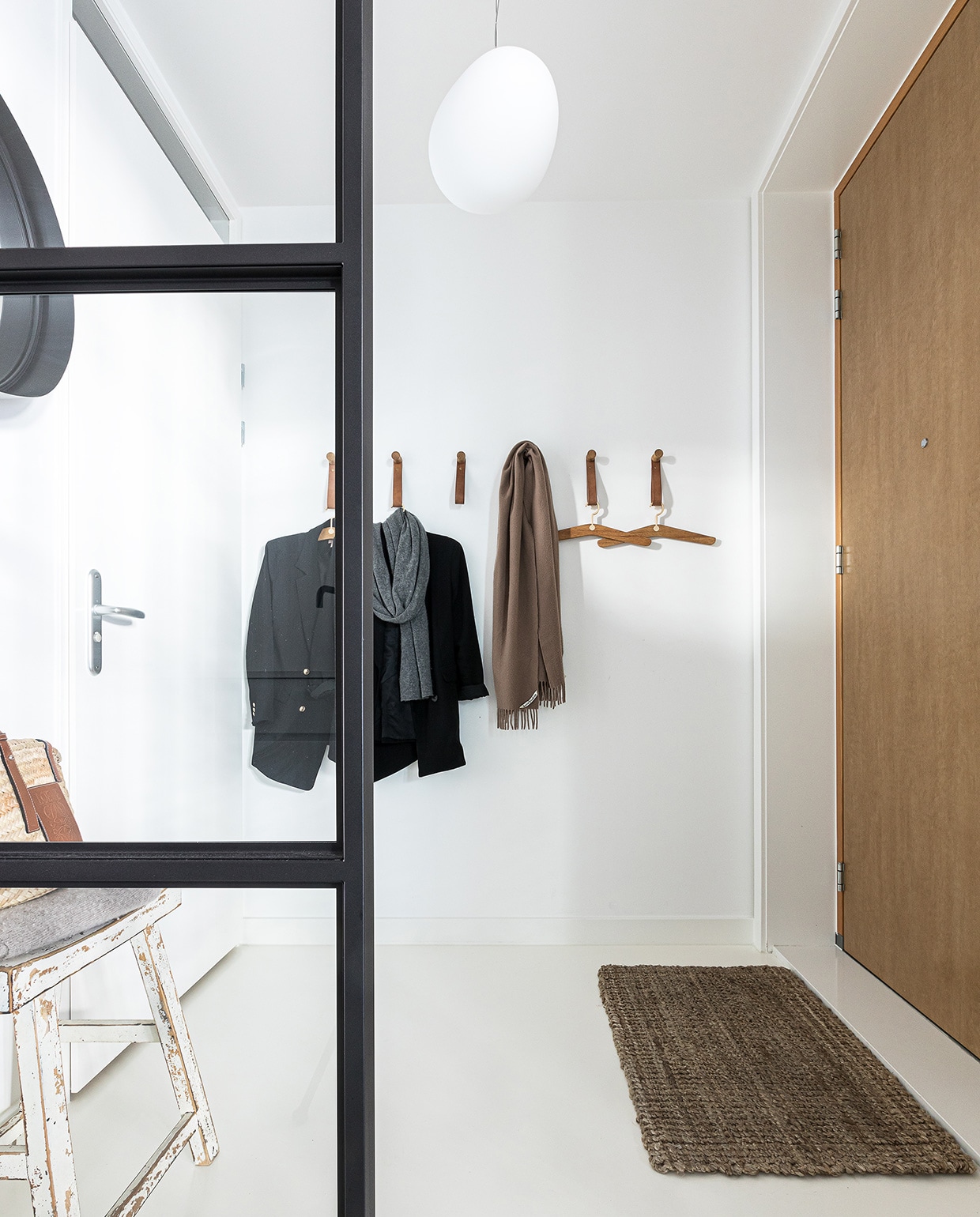 Hanglamp Gregg van Foscarini. Stalen deur Preferro. Houten kruk antiekwinkel. Rieten tas Loewe. Leren kapstok, kledinghangers Bolia. Spiegel Maisons du Monde. Deurmat Home Delight.