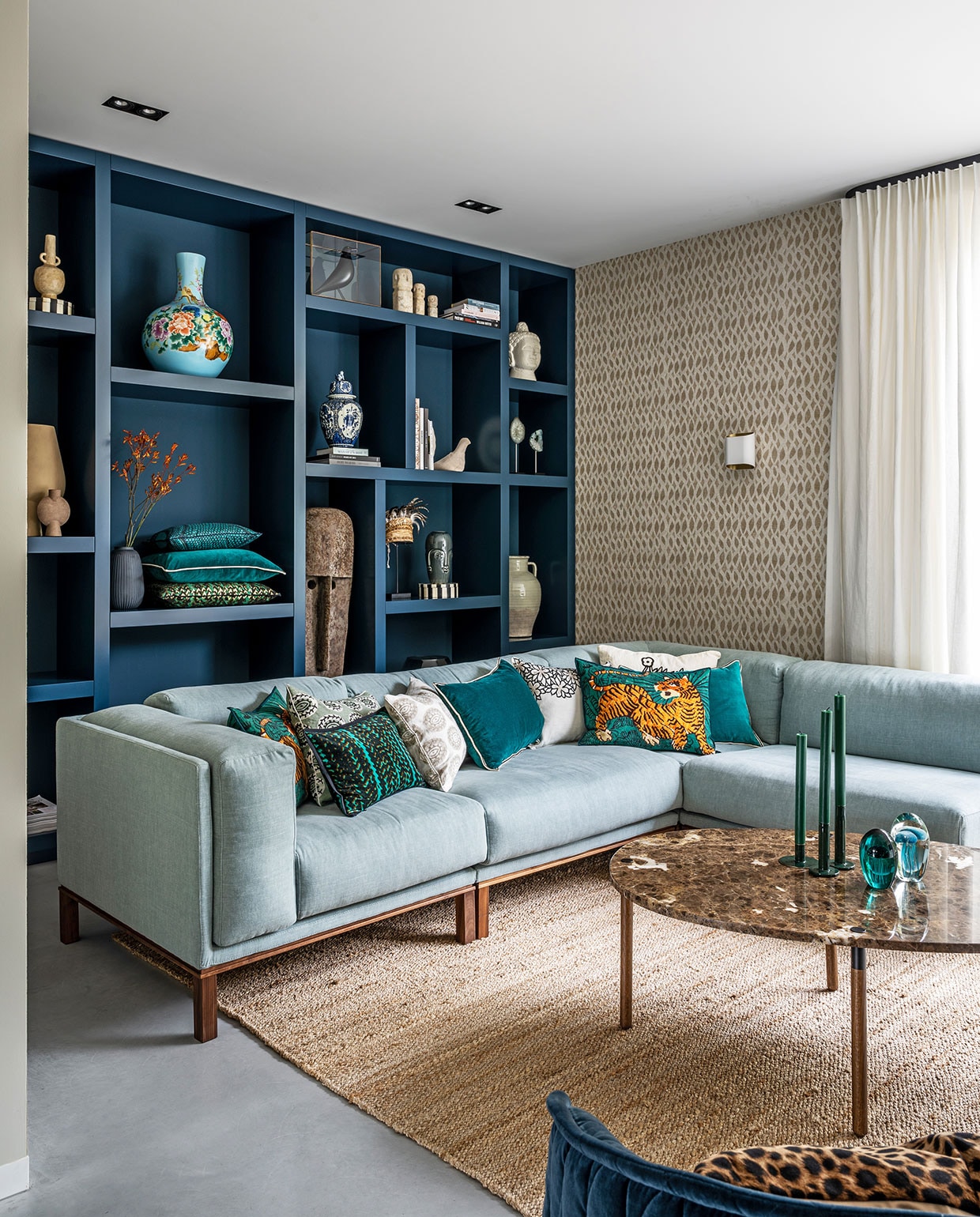 Vakkenkast Maek Meubels in kleur Hague Blue van Farrow &amp; Ball, behang Perles Topaze van Élitis, linnen gordijnen van Interstil, bank en salontafel Bolia, vloerkleed TineK Home, kussens Élitis, kaarsen Sissy-Boy, geëmailleerde groene kaarsenstandaards Tica. In kast: lichtblauwe vaas Pols Potten, zandkleurige vaas en bruine vaas Casa Gitane, witte vogel Pigeon van Christien Meindertsma, zwarte vogel House Bird Vitra Eames, witte figuren rechtsboven Stone Man Dinor en hoed via Loods 5, overig privebezit.