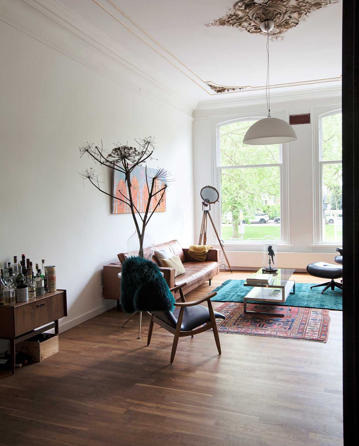 Vintage drankenkabinet, houten stoel met blauwe bekleding en salontafel Bestwelhip. Groene vacht en bijzettafel met driepoot Kieveen. Bruine bank Albufera van Lederland, kussens de Bijenkorf, gele plaid De Groene Lantaarn. Schilderij boven de bank van Merijn Kavelaars. Staande lamp 360Volt. Turquoise kleed Kieveen, Perzisch kleed van oma. Glazen stolp met vogel herkomst onbekend.