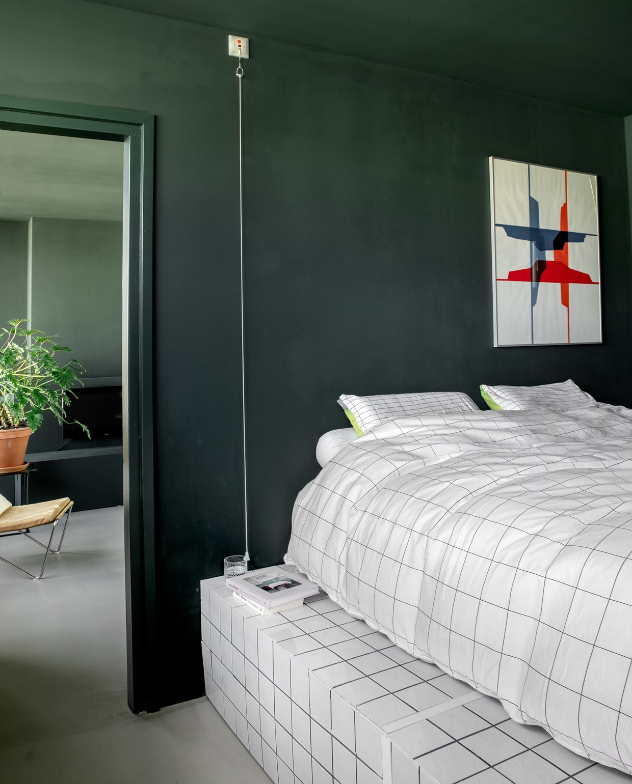 Doorkijkje naar de woonkamer vanuit de slaapkamer. Het plateau waarop het bed staat, werd gebouwd van mdf en vervolgens betegeld. Chantal: ‘Die donkere kleur op muren en plafond maakt het extra knus. Deze kamer voelt als een soort mega bedstee.’ Vintage Bachelor Chair van Verner Panton via Niet Nieuw Design. Plant en terracottapot Intratuin. Witte tegels 10×10 cm Hornbach. Boek Little Escapes via bartsboekje.com.