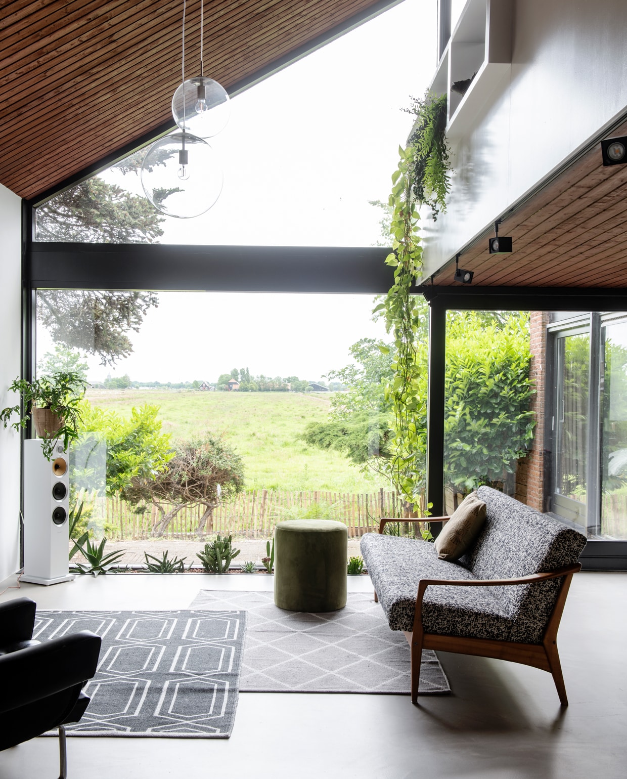 De woonkamer met uitzicht op de polder. Vloerkleden H&amp;M, vintage bank Decennia Design, groene poef van Riverdale, witte speaker Bowers &amp; Wilkins, bollampen van Rietveld Licht &amp; Wonen, spots Altra Dice via slv.com. De oorspronkelijke convectorput doet nu dienst als XL plantenbak.