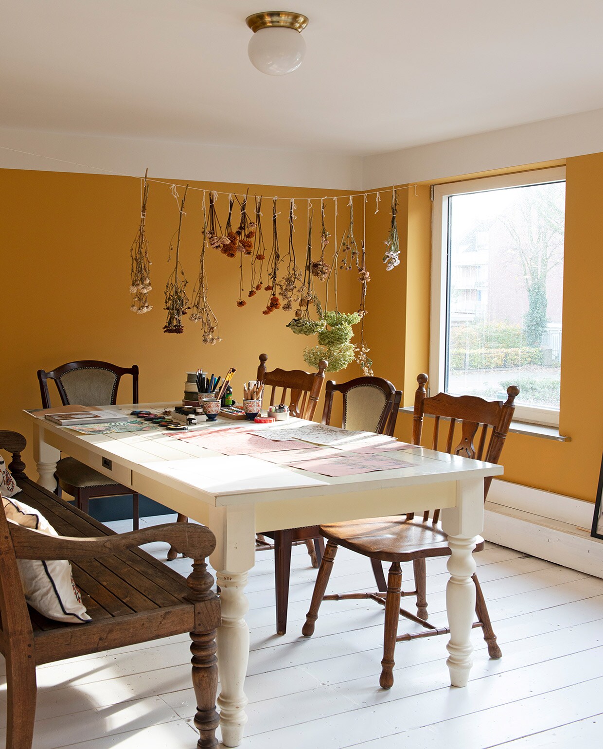 Muurverf India Yellow van Farrow &amp; Ball. Verf plafond Slaked Lime van Little Greene. Vloer origineel. Plafondlamp Ikea. Bloemen zelf gedroogd. Tuinbank gekregen, kussens Ikea. Tafel Marktplaats. Gestoffeerde stoelen kringloopwinkel. Houten stoelen en bekers gekregen.