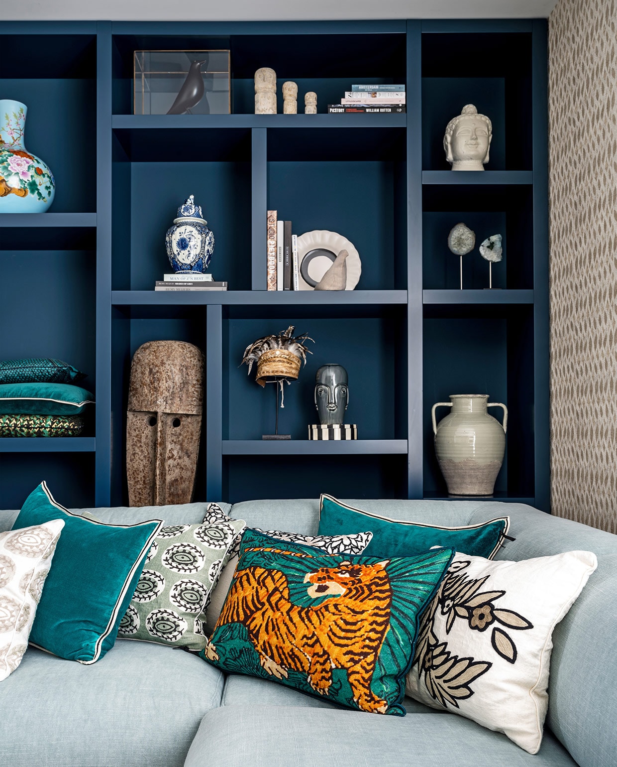 Vakkenkast gemaakt door Maek Meubels Heerhugowaard geschilderd in de kleur Hague Blue van Farrow &amp; Ball, behang Perles Topaze van Élitis bank Bolia, kussens Élitis. In kast: lichtblauwe vaas Pols Potten, zandkleurige vaas en kleine bruine vaas Casa Gitane, witte vogel Pigeon van Christien Meindertsma, zwarte vogel House Bird Vitra Eames, witte figuren rechtsboven Stone Man Dinor en hoed op standaard Bali via Loods 5, overige items souvenirs en cadeaus.