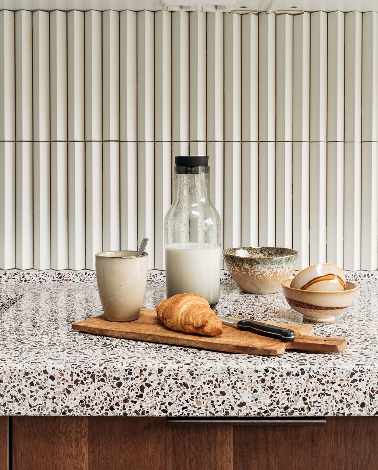 Basis keuken Ikea, fronten in kleur Smoked Walnut (onder) en Kasjmiergrijs via Frontz, terrazzo blad Rosa Bian Terrazzowerken. Wandtegel Rombini triangle small white van Mutina. Plank Action, mokken Xenos, karaf Hema. Verf muur en plafond R0.10.60 van Sikkens. Stoelen Little Tulip van Artifort via Marktplaats. Tas Rough Studios. Vloer Cem-Line Micro van Loos Betonvloeren.