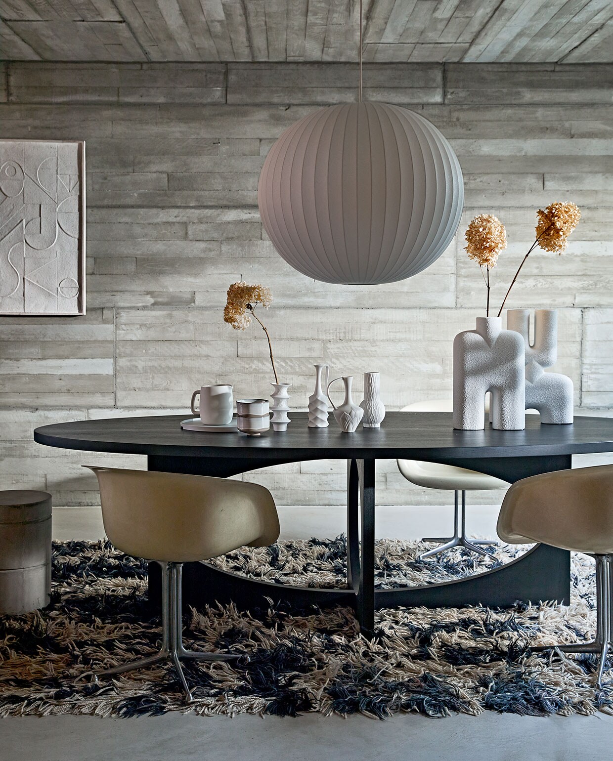 Zwartgebeitste massief eikenhouten ovale tafel van ontwerper Sarah Trenite, 260x115x75 cm (lxbxh), prijs op aanvraag (). Vintage La Fonda kuipstoel van Eames voor Herman Miller, prijs op aanvraag (pietjonker.nl). Grote witte vazen Cobra Bubble tall € 120,- en Double Bubble tall € 135,- van 101CPH (edha-interieur.nl). Schilderij met reliëf, 100×70 cm € 199,- (urbannatureculture.nl). Kleine vazen van HKliving, set van 4 € 129,95, waterkan keramiek van ontwerper Vincent van Duysen voor Serax, 1,5 liter € 25,-, hanglamp Nelson Ball Bubble van Hay, 68 cm (Ø) € 607,99, dienblad van Zone Denmark, 35 cm (Ø) € 50,-, kommen Merci n°1 van Serax, 12,5 cm (Ø) per stuk € 16,65 (vtwonen.nl). Betonnen kruk van Marie Michielssen, 30×45,5 cm (Øxh) € 103,- (serax.com). Gedroogde hortensia’s (edenique.nl). Soortgelijk vintage vloerkleed (vintagehoarder.com).