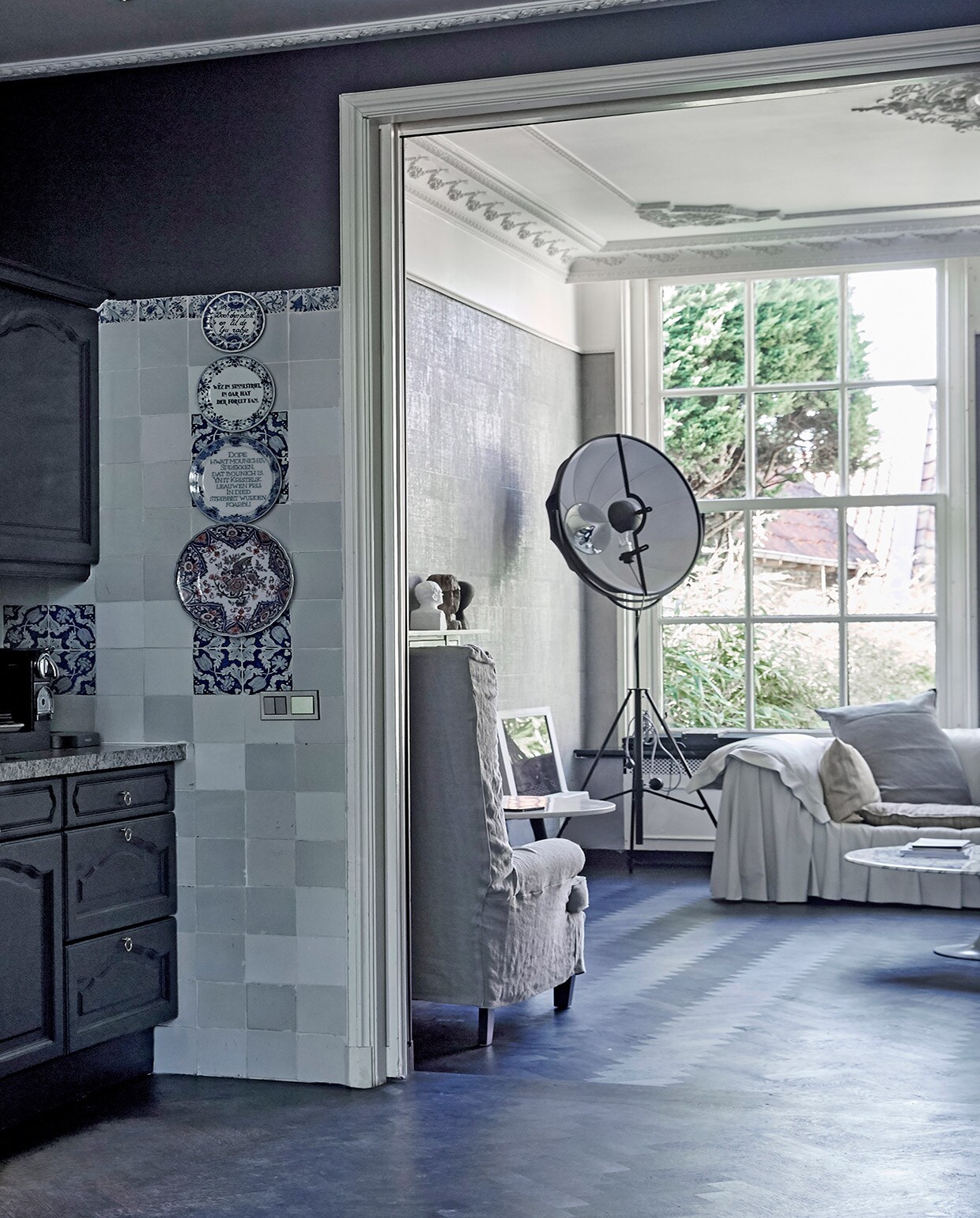 Keuken donkerblauw geschilderd. Tegels op achterwand originele Harlinger tegels. Borden met Friese spreuken aan muur vintage. Doorkijkje naar de achterkamer: stalamp Fortuny van Pallucco, leren bank Bohémien van Busnelli, kussens op bank gemaakt van stoffen van Emery &amp; Cie. Witte tv Bang &amp; Olufsen. Witte buste van een winkel die niet meer bestaat, antieke houten buste uit India, zwart hoofd van GMC uit Zuid-Afrika. Lage marmeren tafel Knoll, hoge marmeren tafel Tom Dixon.