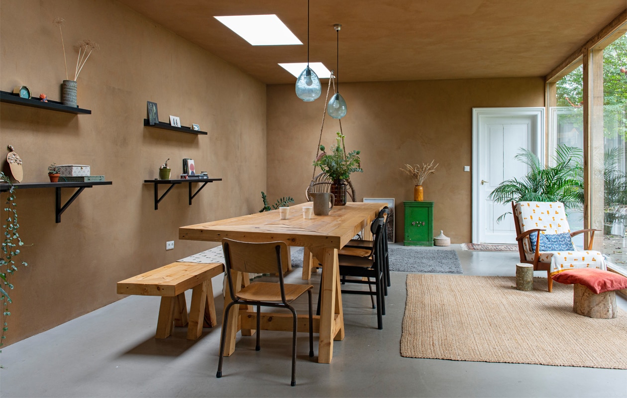 Wanden en plafond van onbehandeld leemstuc, vloer van beton. Muurplanken, vloerkleden, stoelen Ikea, hanglampen Sissy-Boy. Tafel en bank gemaakt door Joost. Vintage schoolstoel, schenkkan en vazen kringloop, kluis en fauteuil vintage vondsten. Rieten mand Wereldwinkel. Deurmat vintage, deken en kussen op fauteuil Ted &amp; Tone. Rood kussen Dille &amp; Kamille. Bijzettafel en kruk zelfgemaakt van boomstammen uit de tuin.