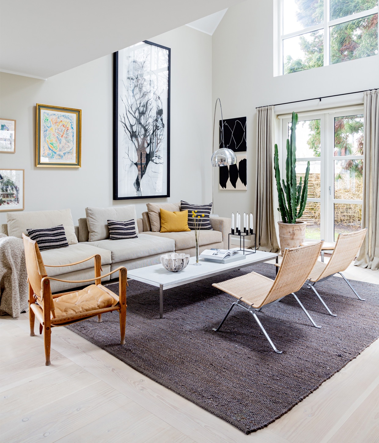 Muurverf Shaded White van Farrow &amp; Ball. Bank Eilersen. Kussens Tapet-Cafe. Koffietafel PK62 en rotan stoelen PK22 van Poul Kjærholm voor Fritz Hansen. Leren stoel Kaare Klint voor Carl Hansen &amp; Son. Vloerlamp Arco van Flos. Schaal gemaakt door een vriendin. Kaarsenstandaard Mogens Lassen. Schilderijen van links naar rechts Carsten Dahl (twee maal), Eigill Jacobsen, Cathrine Raben Davidsen. Grafische kunstwerken Ulla Pedersen. Vloerkleed Tine K Home.
