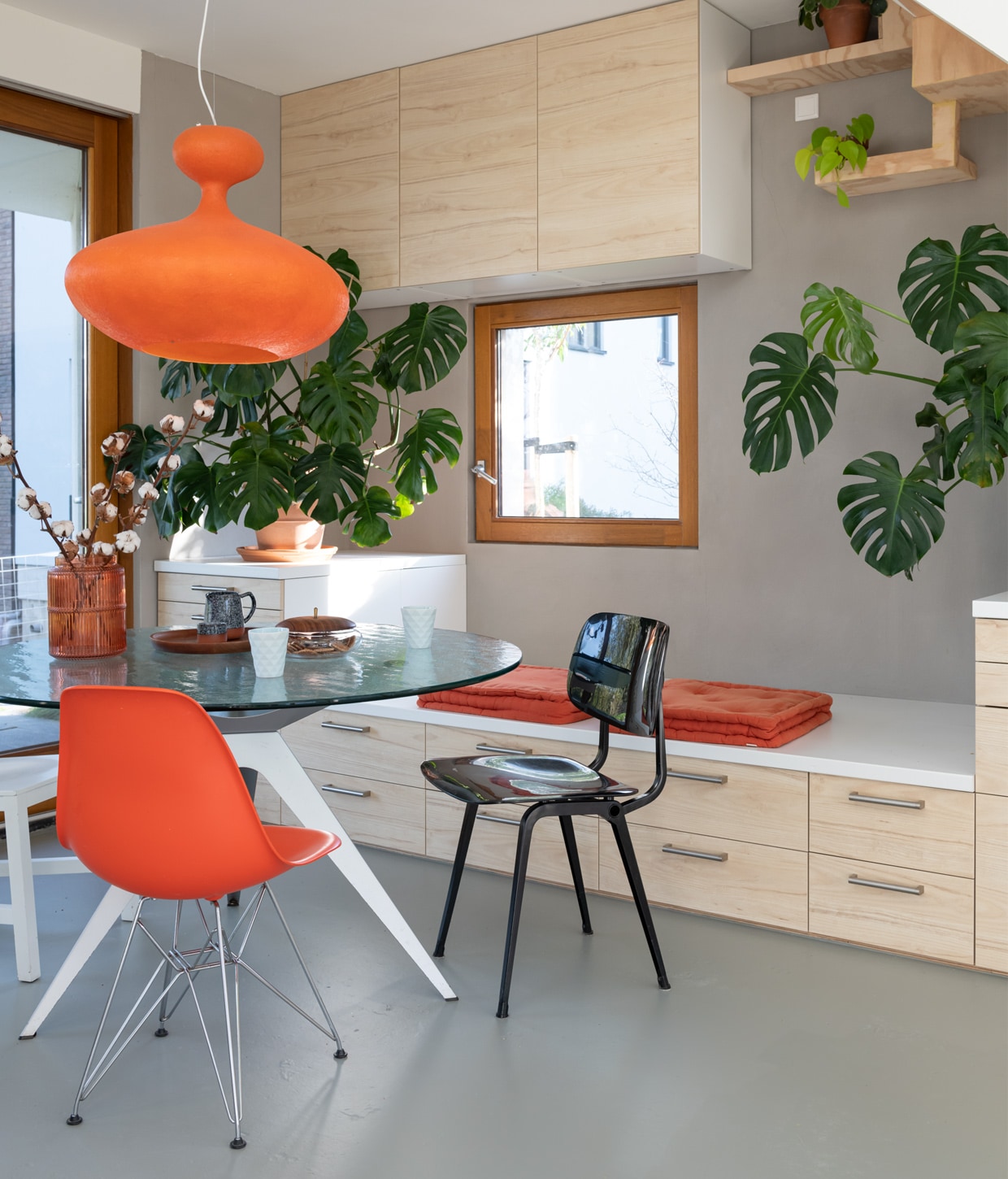 Zandcementdekvloer met vloercoating, trap en houten muren van vurenhout en radiplex, eettafel Ben Hoek Meubelarchitectuur, lamp Kundalini E.T.A. Sat, oranje stoel Charles Ray Eames, zwarte stoel Revolt van Friso Kramer, witte stoel Joep van Lieshout, oranje vaas Kwantum, dienblad H&amp;M Home, kan Søstrene Grene, lichtblauwe mokken kringloop, koektrommel met houten deksel en grijze aardewerken pot vintage, oranje palletkussens tuincentrum, terracotta potten Intratuin en Ikea, handgrepen lades Ikea.
