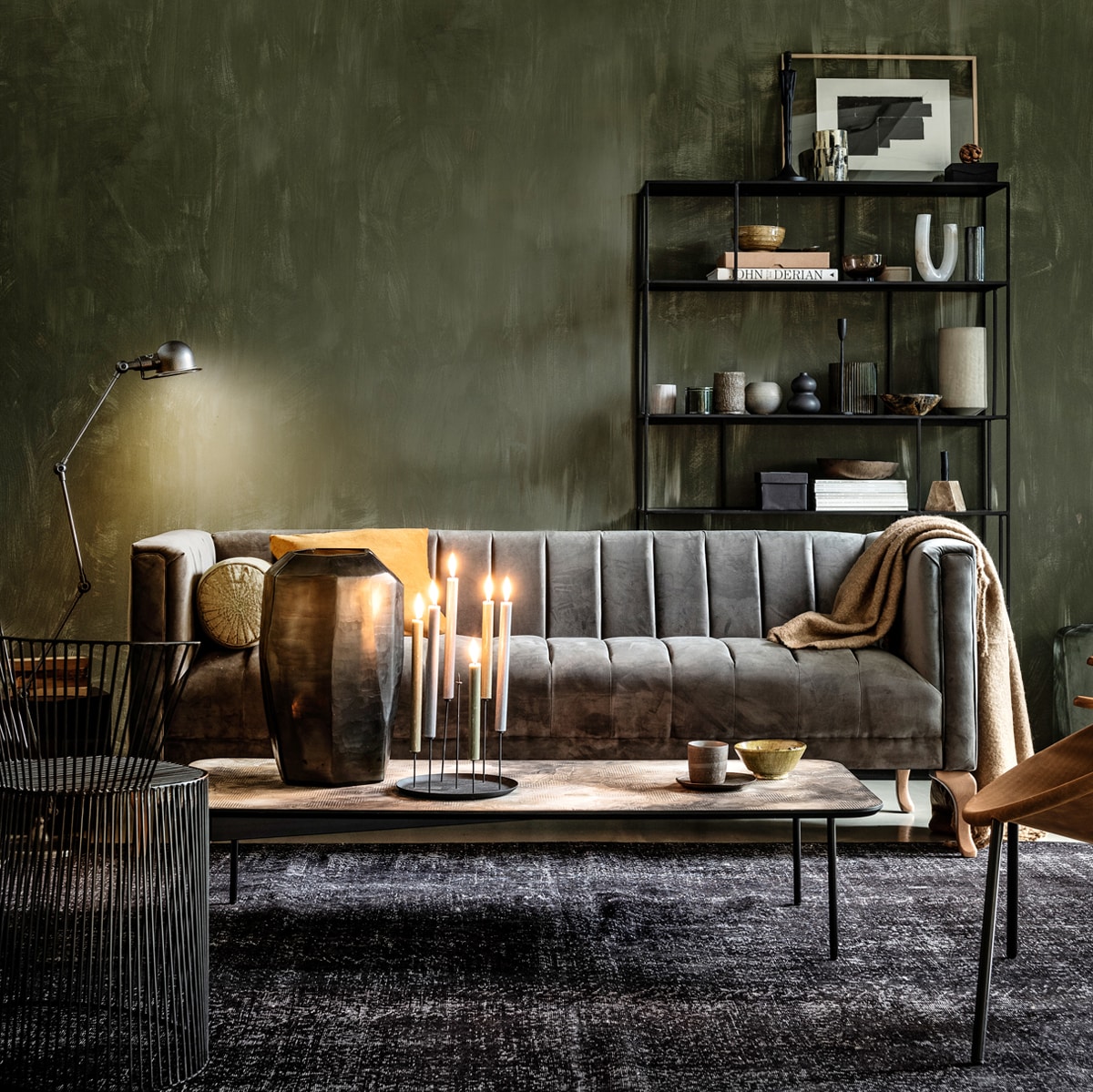 Grijze zetel met velours bekleding 205x77x80 cm (bxhxd) (rivieramaison.nl). Verf op wand Fresco kalkverf in kleur Belgian Wilderness, (pure-original.nl). Metalen 4-arm vloerlamp van Jieldé (vdle.nl). Zwarte metalen kast 110x30x180 cm (lxbxh) (polspotten.nl). Groen rolkussen, mosterdgele kom (loods5.nl). Geel velvet kussen, beige wolmix plaid (hm.com). Salontafel, glazen vaas (homestede.nl). Zwarte kandelaar (klevering.nl). Beker, schotel (thingsilike- thingsilove.com). Draadstalen stoel (covanderhorst.nl). Fauteuil met suède zitting (goeds.nl). Zwart vintage vloerkleed 304×172 cm (lxb) (rozenkelim.nl). Kast van boven naar beneden, van links naar rechts: zwart beeld, groene pot met deksel, zwarte metalen kandelaar, bronzen schaal (thingsilikethingsilove). Houten lijst, schaal van hoorn (letoile.store). Zwart-witte waxinelichthouder, mosterdgele kom, witte marmeren kandelaar, marmeren blokkandelaar (loods5.nl). Bruine glazen kom, marmeren pot met deksel, zwart bolvaasje, grijs geribbeld glazen vaas (hm.com). Zachtgrijze vaas (homestede.nl). Overig privébezit.